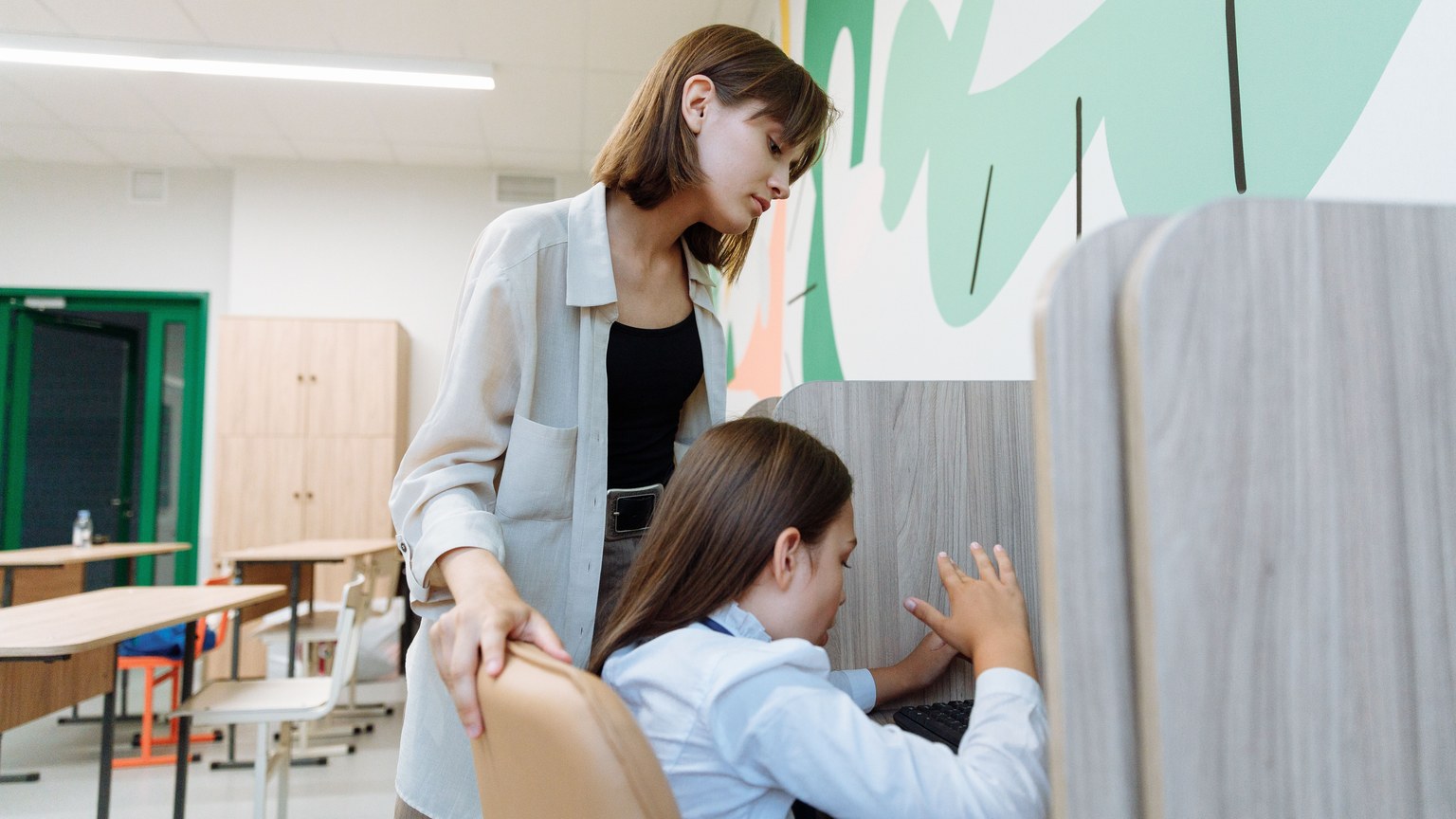 A teacher assists a student working on a computer in a bright, modern classroom setting.