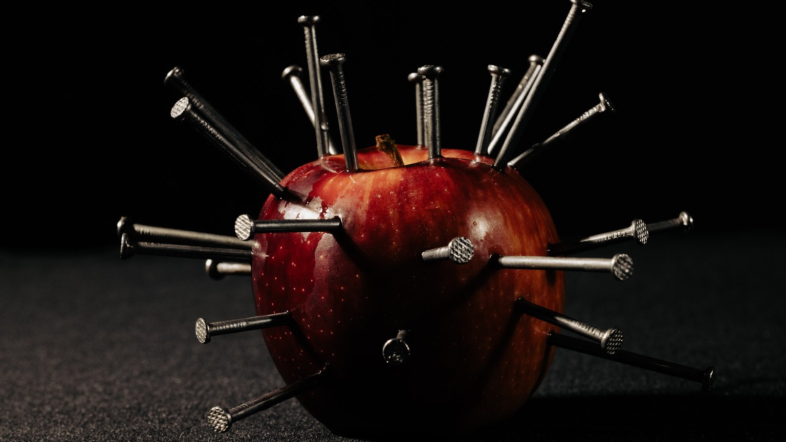 A red apple pierced by multiple nails against a dark background, representing danger.