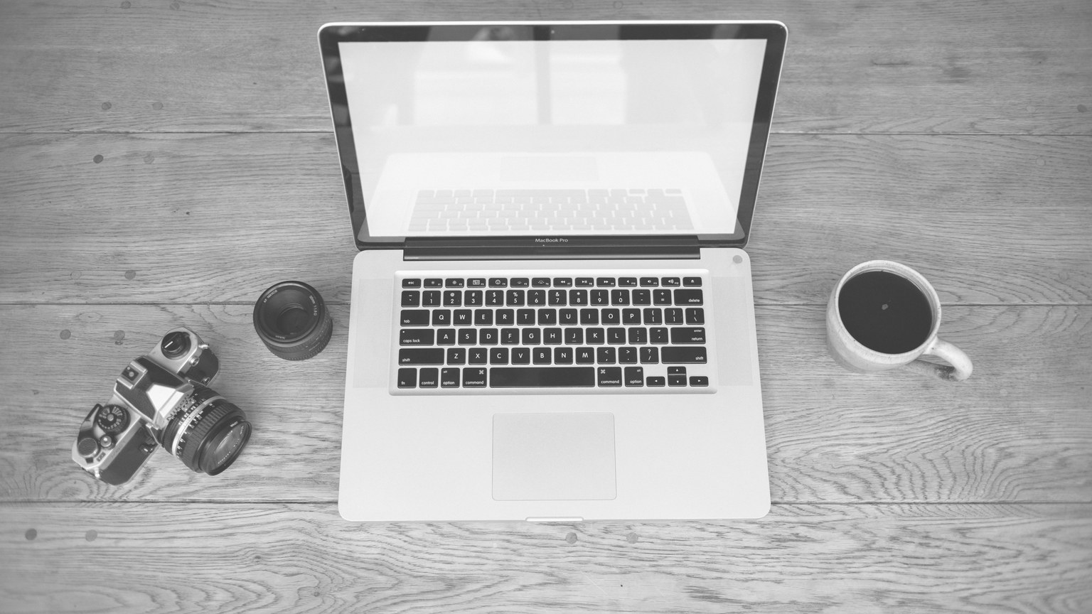 Monochrome image of a laptop, camera, lens, and coffee cup on a wooden desk