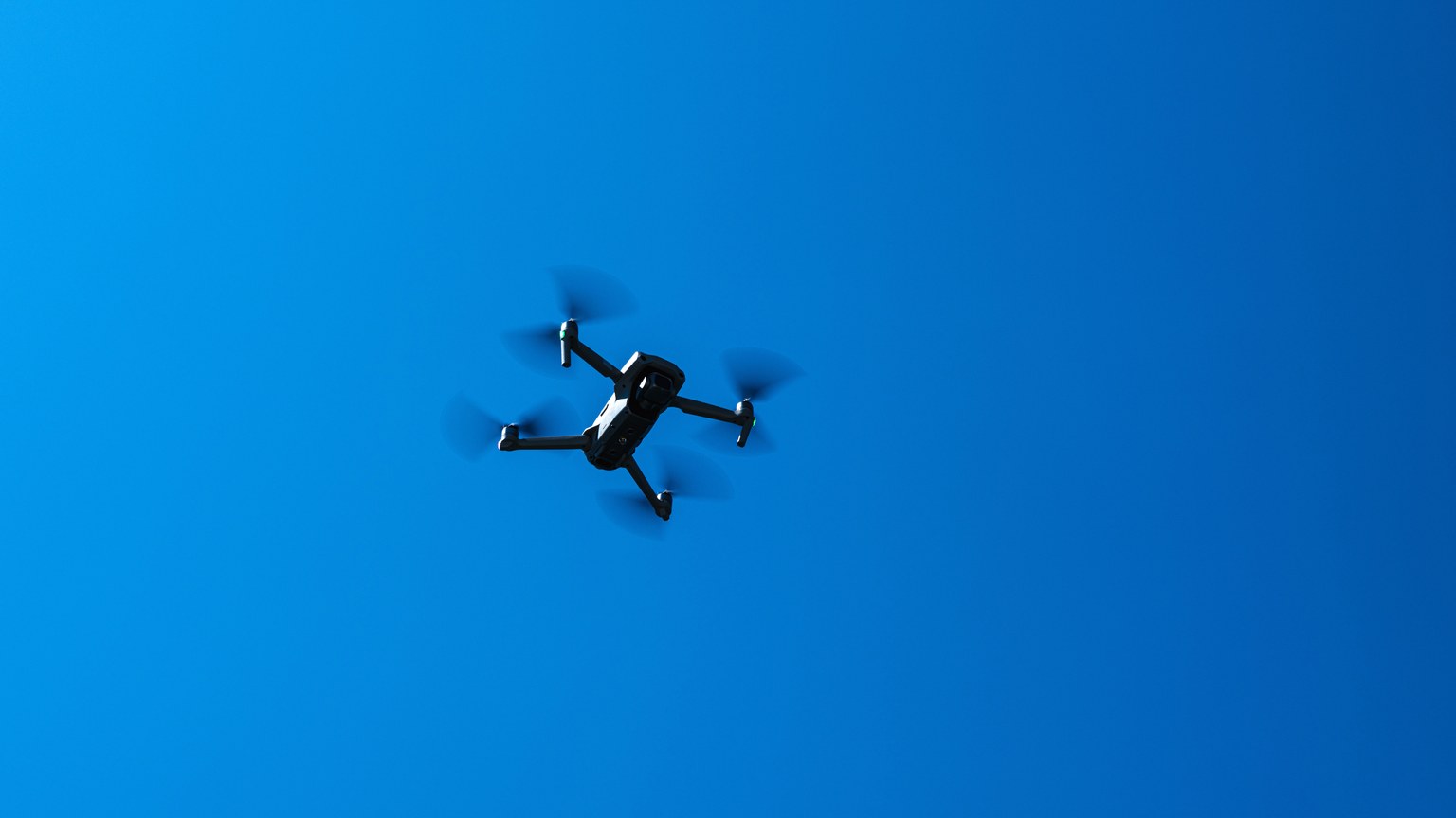 A black drone flying high in a clear blue sky, showcasing modern technology and flight capabilities.