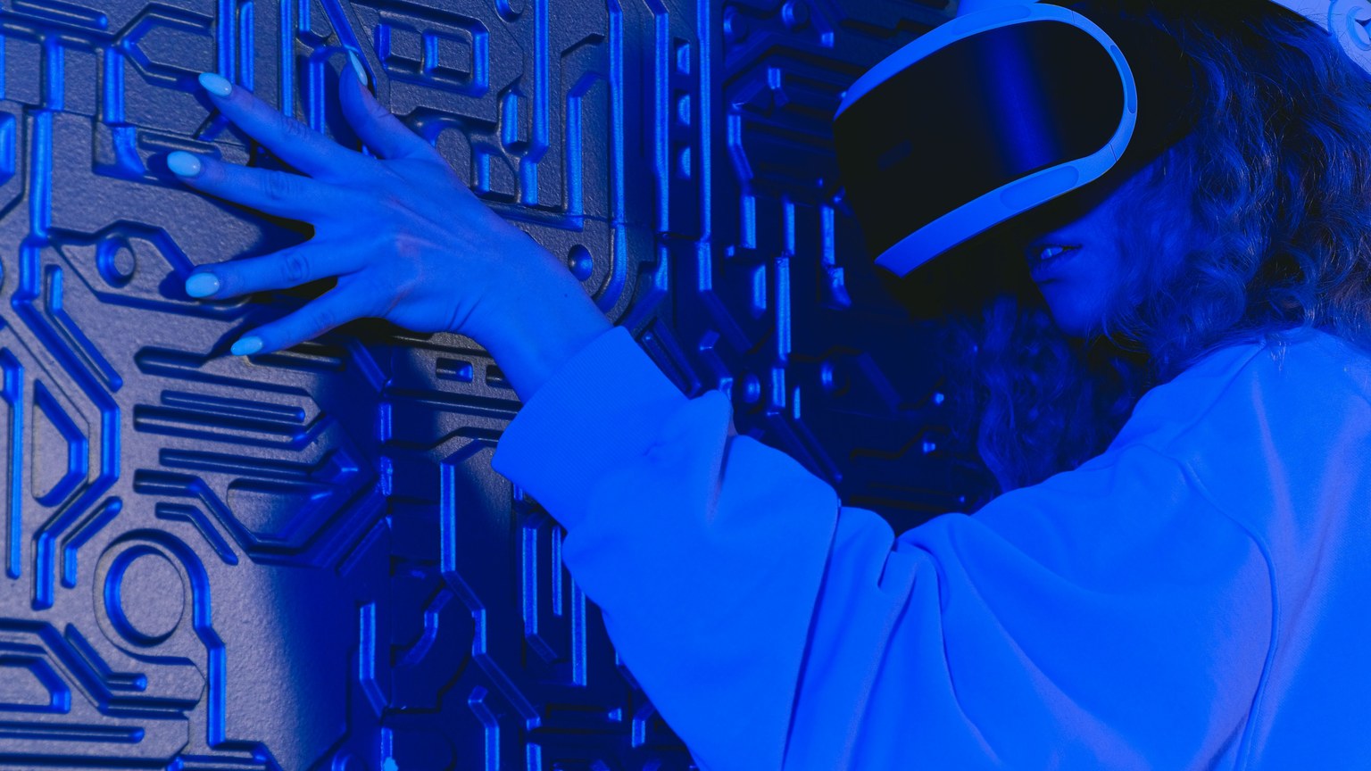 A woman wearing a VR headset touches a textured wall in a blue-lit futuristic environment.