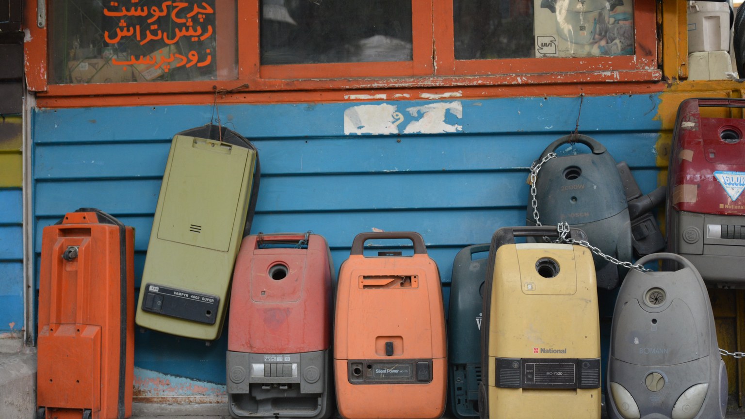 A row of colorful vintage vacuum cleaners displayed against a blue wall in Tehran, Iran.