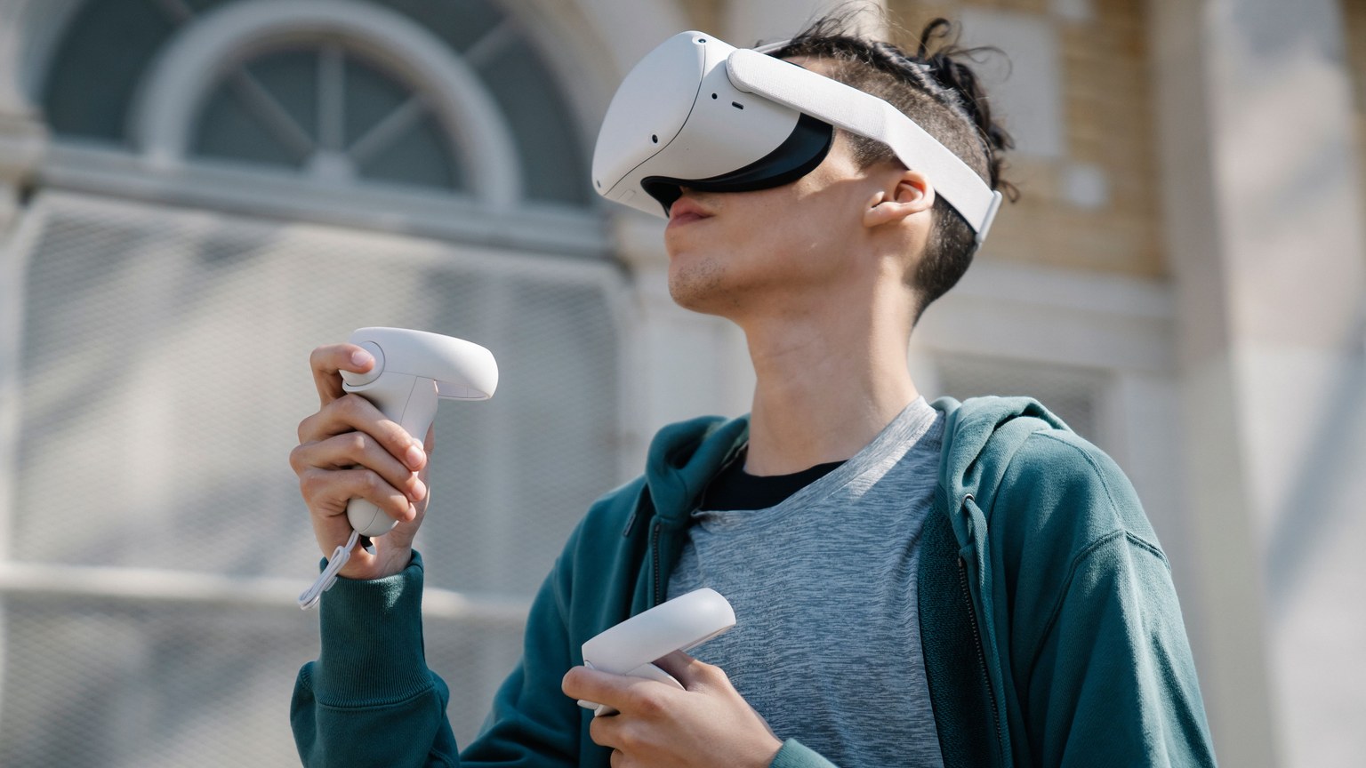 Low angle of young male in blue hoodie using modern wireless virtual reality headset and controllers near building on street