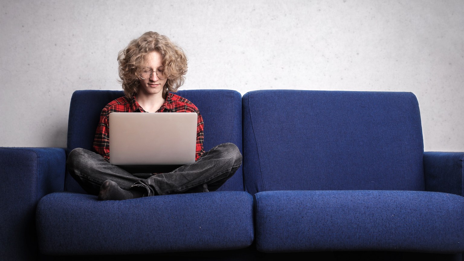 Young adult sitting on blue sofa using laptop, representing remote work lifestyle indoors.