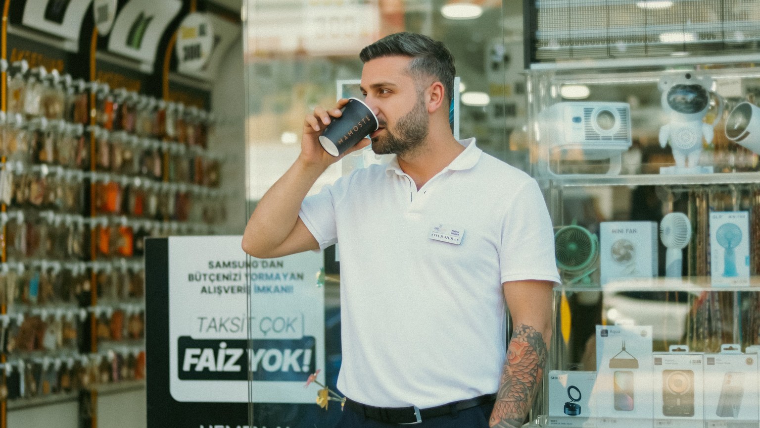 Young man sips coffee while standing in front of an electronics shop, surrounded by gadgets.