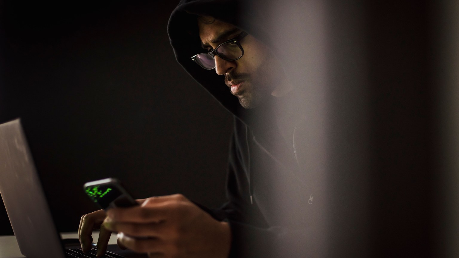 Crop bearded ethnic male computer geek in hoodie chatting on cellphone while typing on netbook in dark room