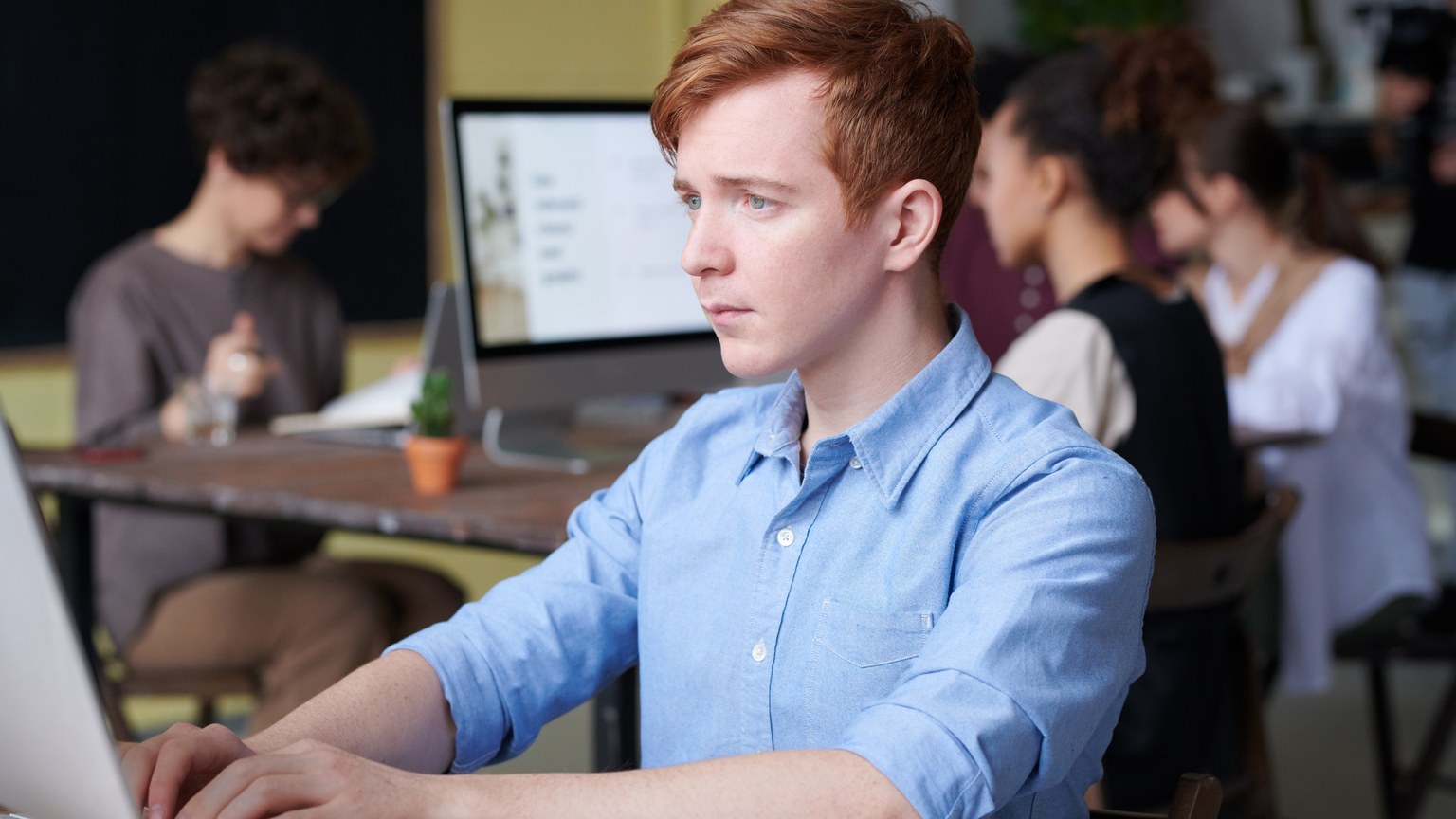 Young professional working intently on a computer in a busy modern office setting, surrounded by colleagues.