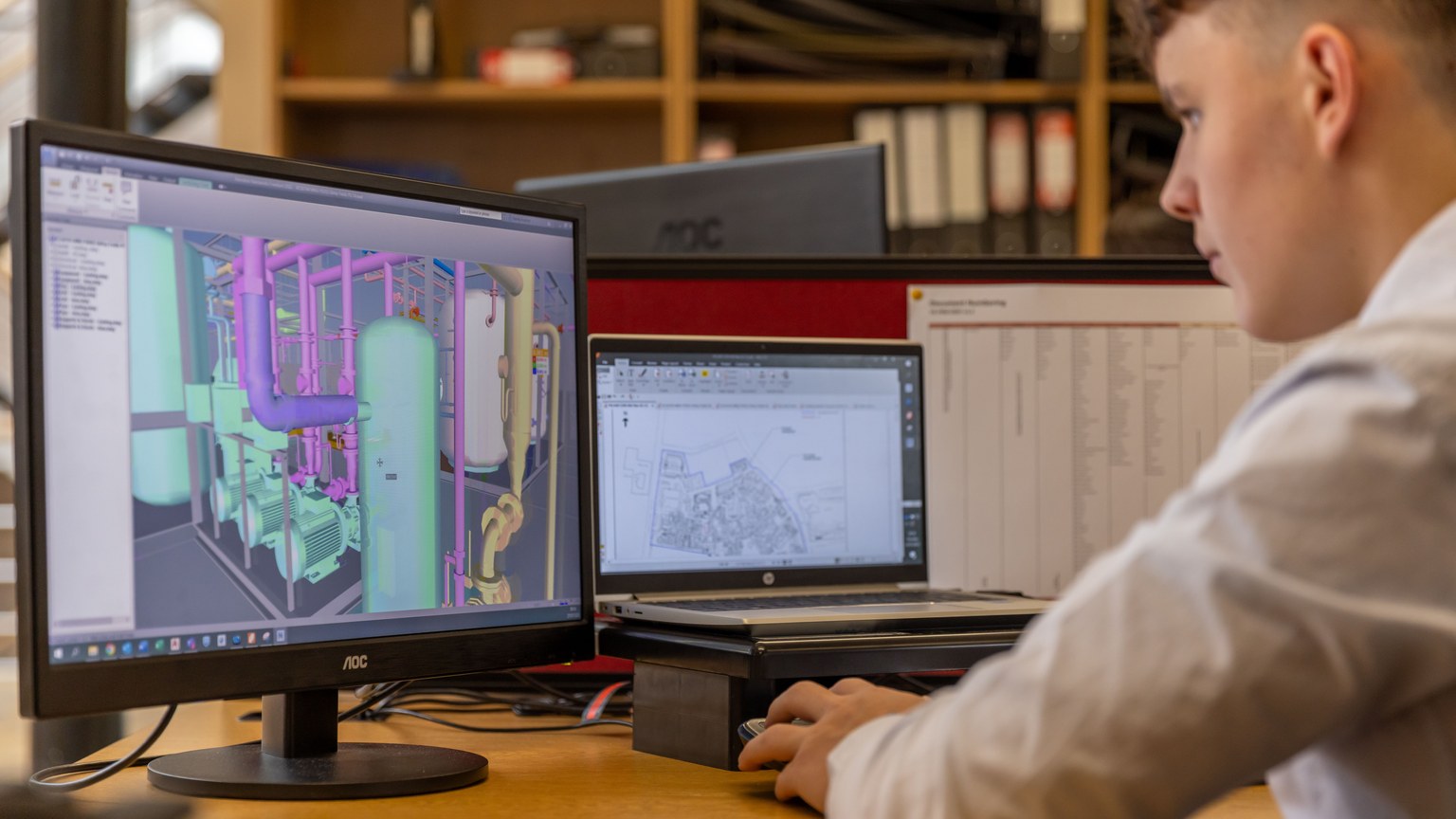 Engineer working with CAD software on dual monitors in an office setting.