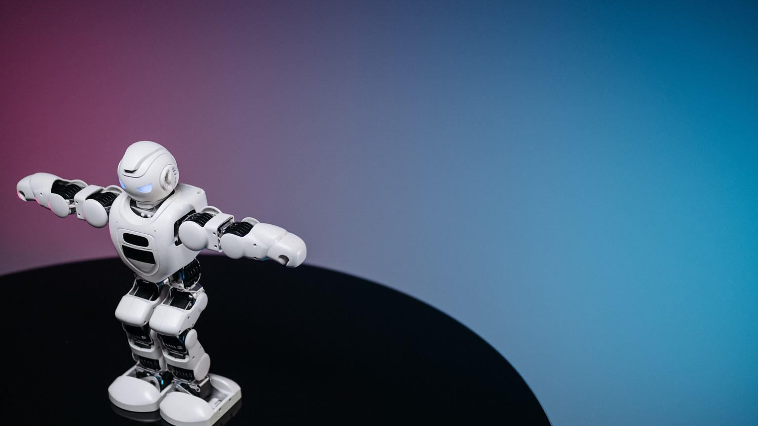 A white humanoid toy robot standing on a reflective black surface in a studio setting with a blue and pink gradient background.