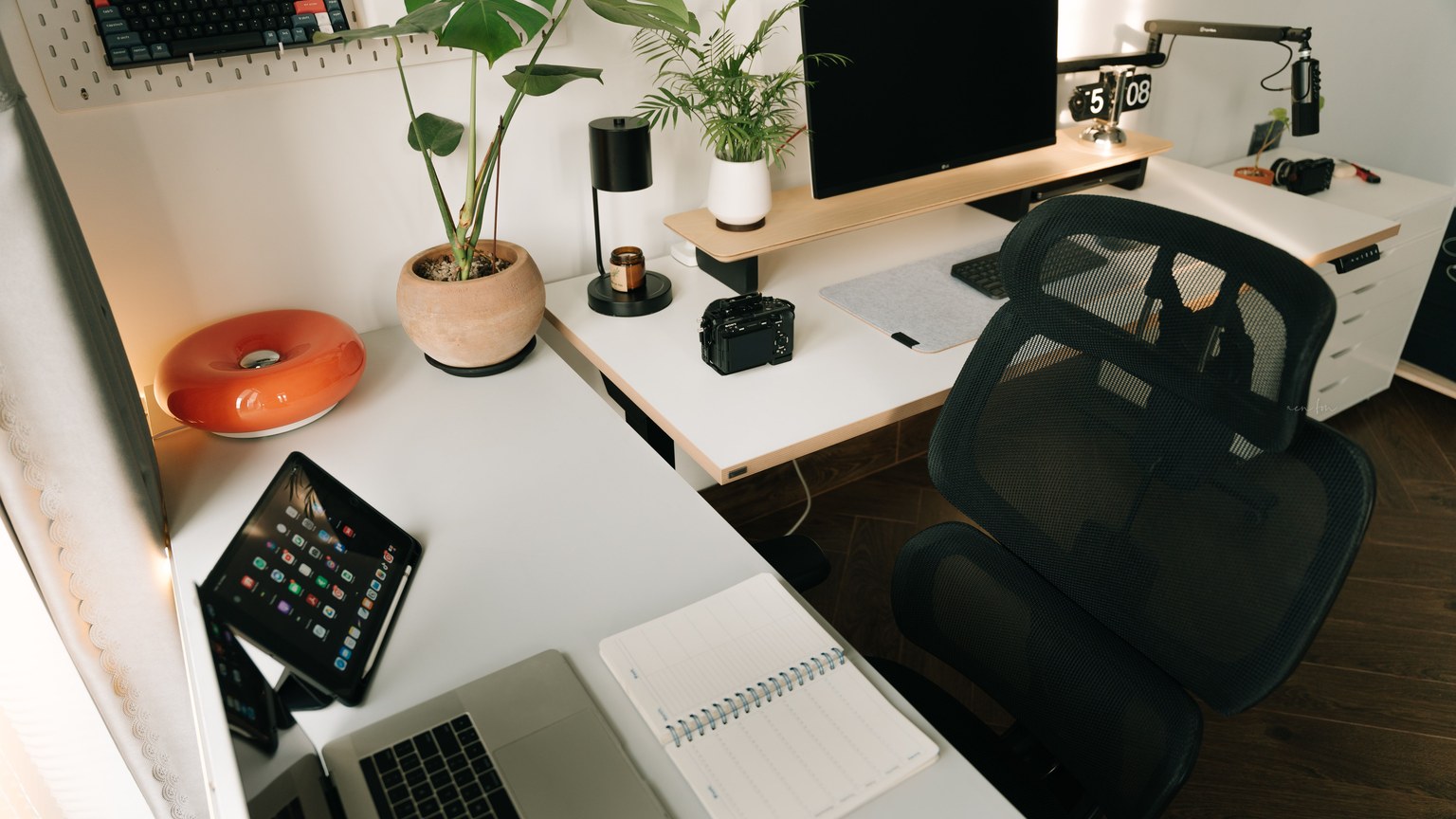 A stylish home office setup featuring tech gadgets, a monitor, a tablet, and decorative plants in a minimalist design.