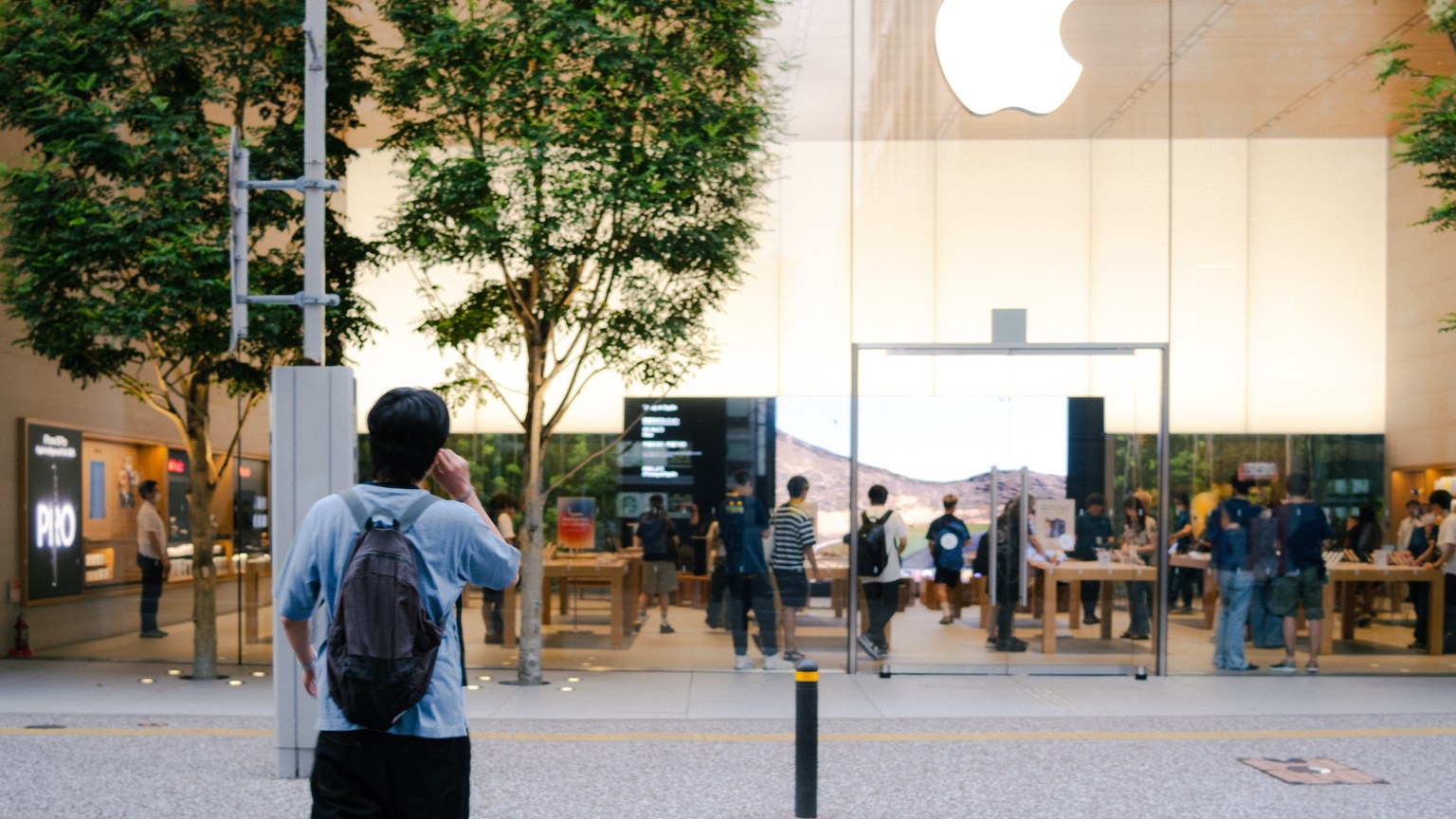 A busy Apple Store entrance with glass facade, trees, and people milling around.