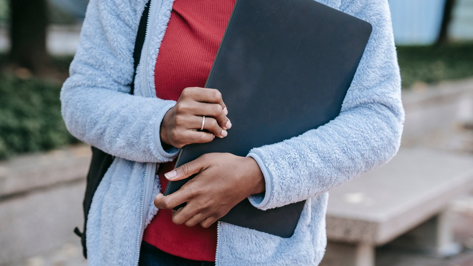 Crop anonymous young African American female student in warm casual clothes and with backpack standing in campus park with laptop in hands