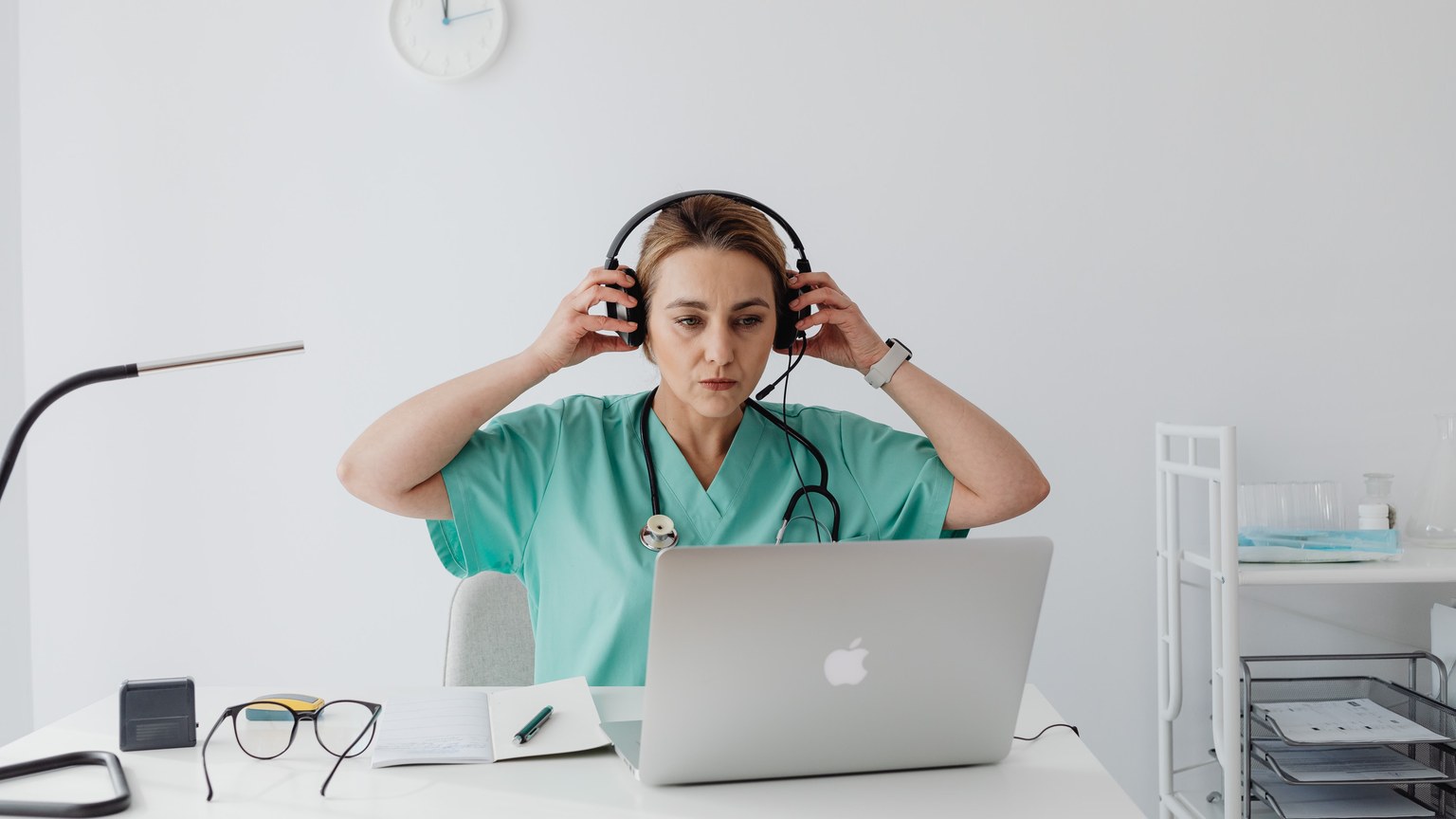 Medical professional in teal scrubs using a laptop and headset, sitting at a desk in an office setting.
