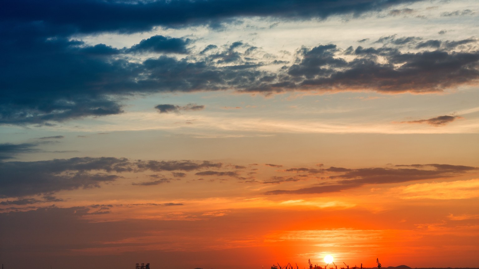 Epic sunset view over the sea with silhouettes of industrial cranes on the horizon.