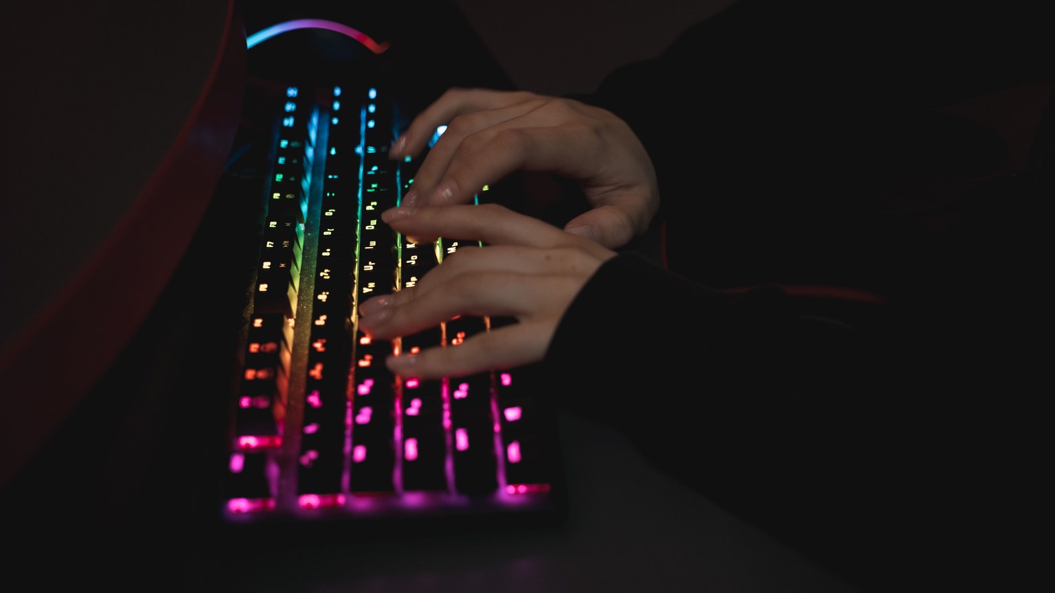 Close-up of hands typing on a colorful RGB backlit mechanical keyboard in a dimly lit environment.