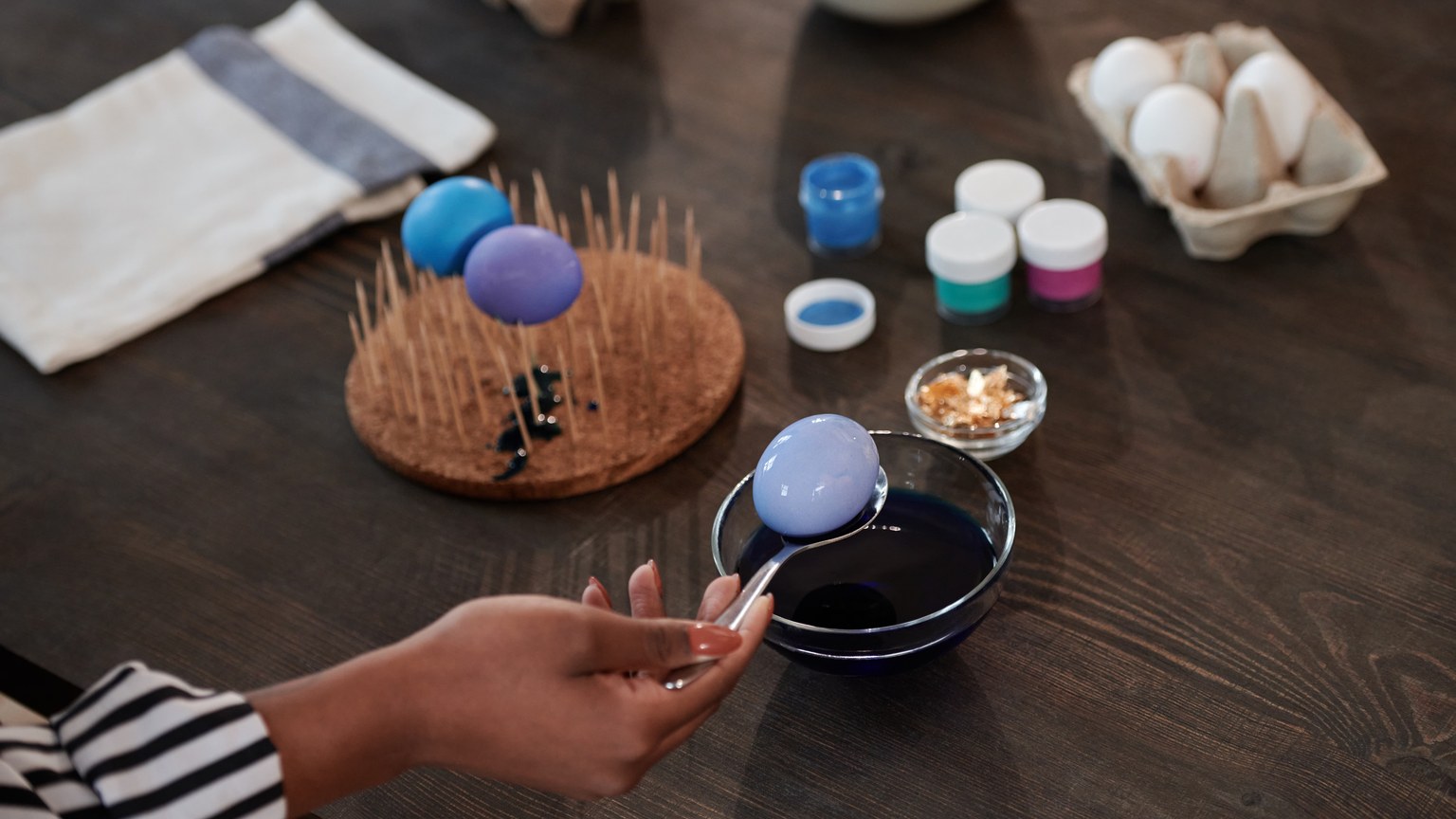 Hand dipping an egg into blue dye, surrounded by painting supplies, for Easter preparations.