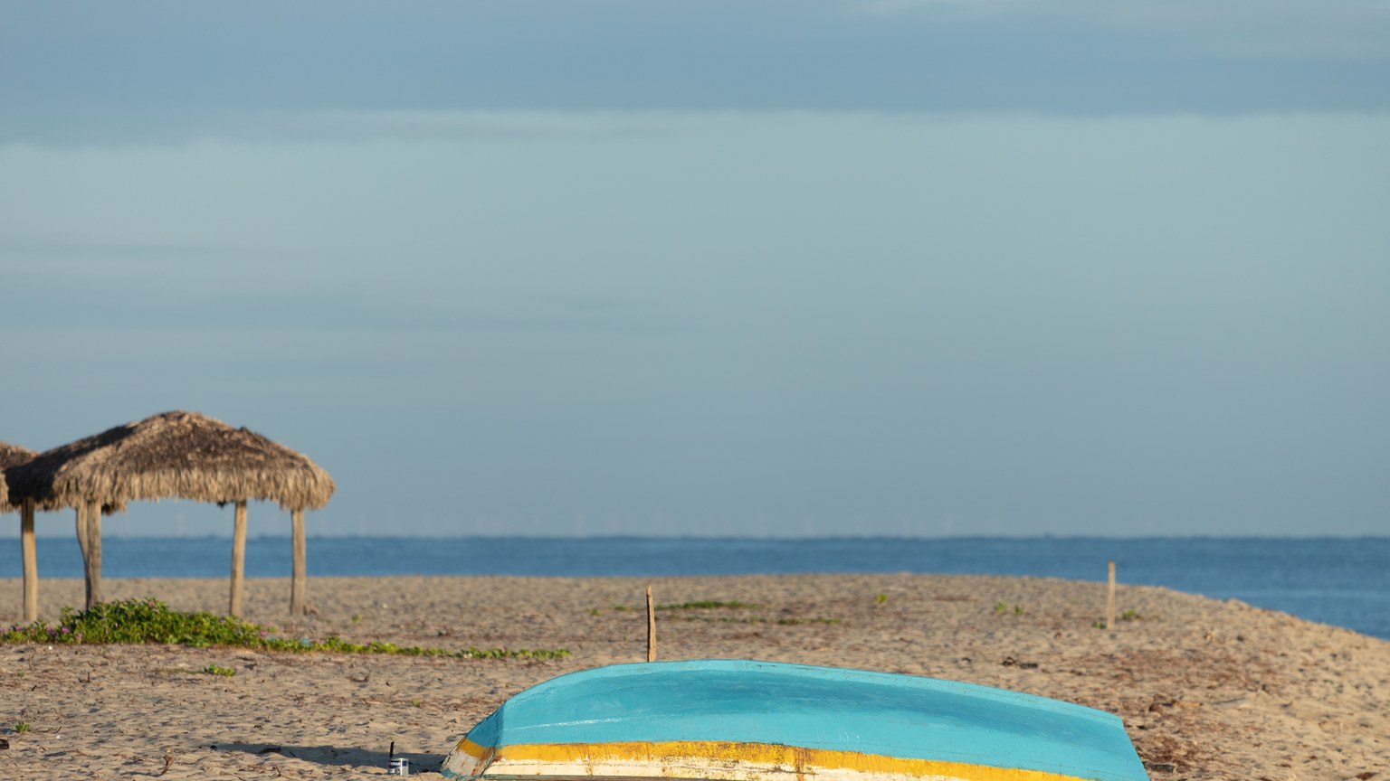 Peaceful beach scene with an upside down boat and straw huts under a blue sky.