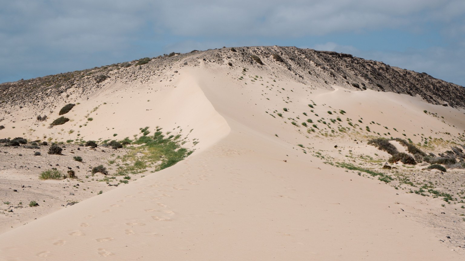 Expansive sand dune with sparse vegetation under a partly cloudy sky.