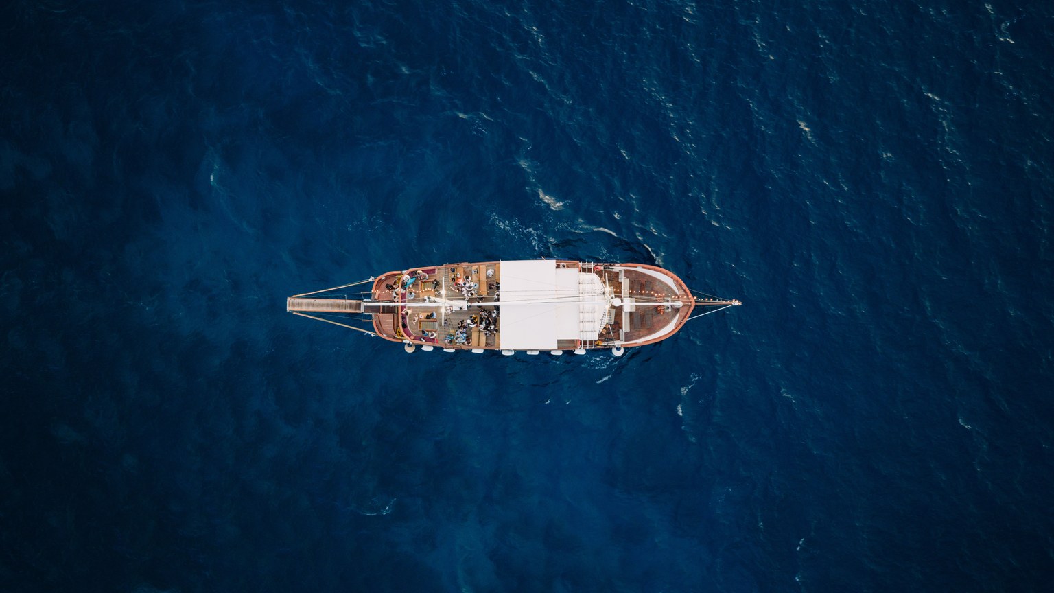A traditional wooden ship sails through the deep blue sea off the coast of Greece, viewed from above.
