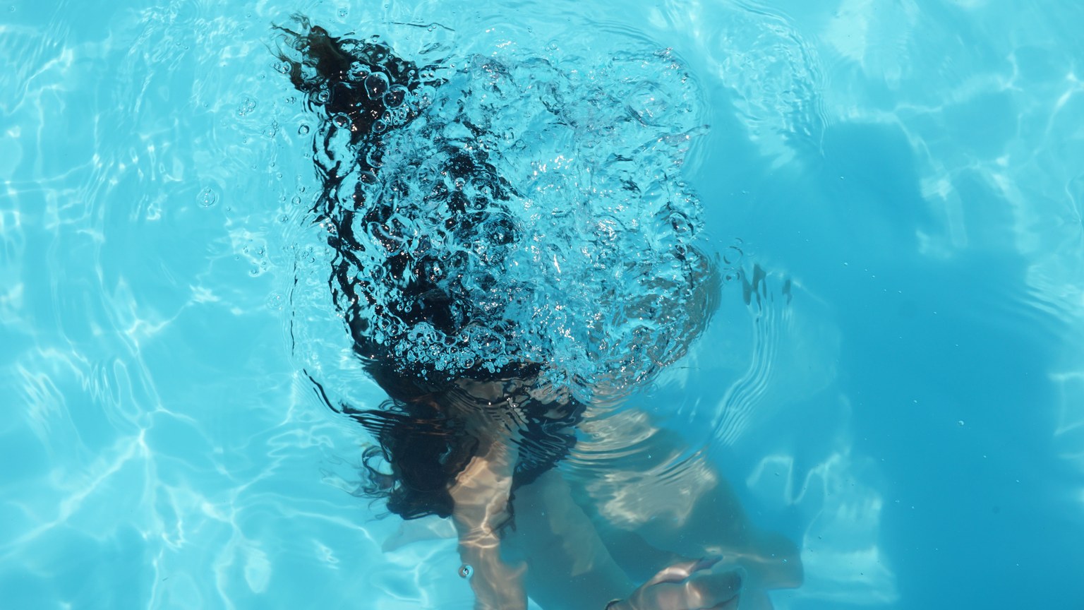 A woman diving underwater in a clear blue swimming pool, captured from above.