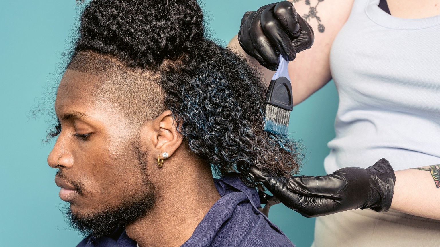 Crop female hairdresser in gloves with brush spreading dye on hair of African American male customer sitting against blue background