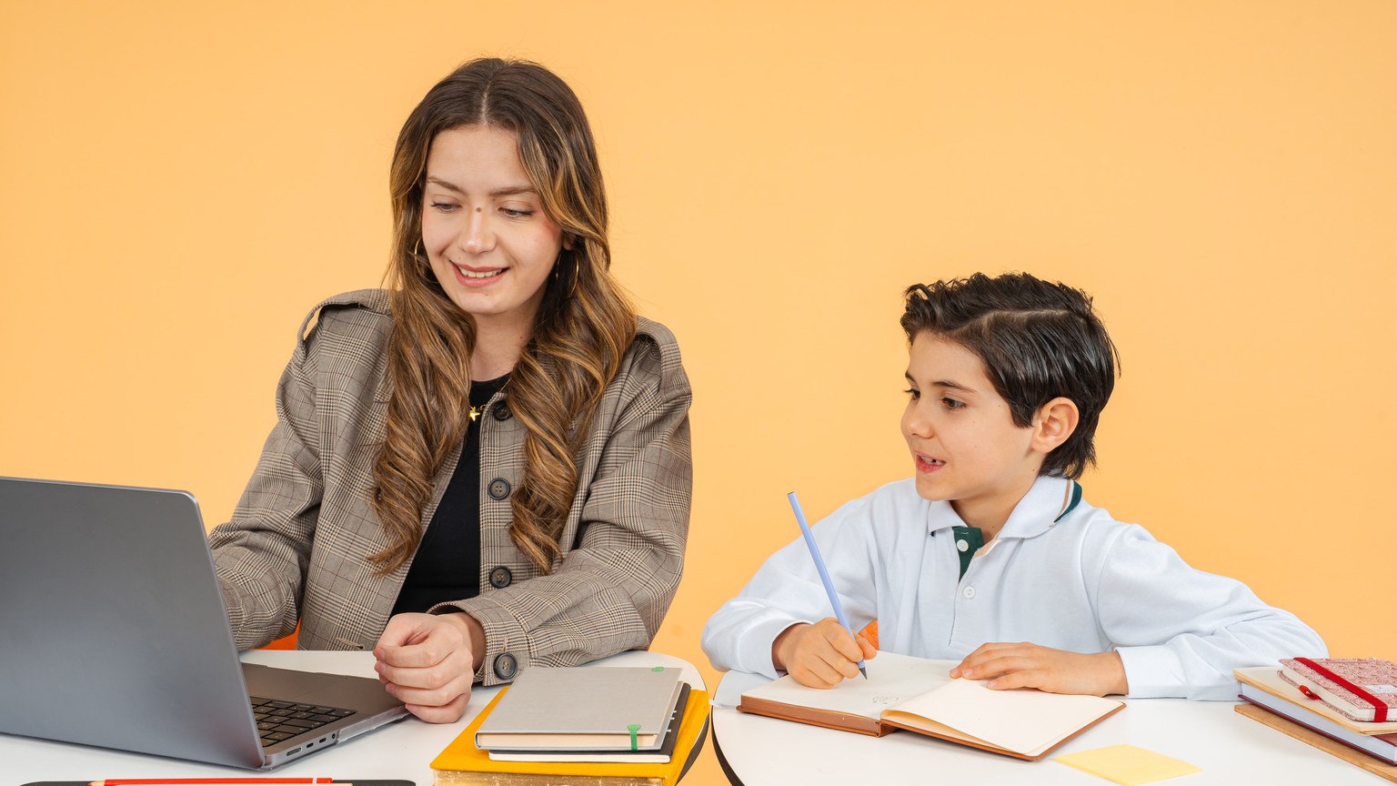 A female teacher helps a young student with his studies using a laptop in a classroom setting.