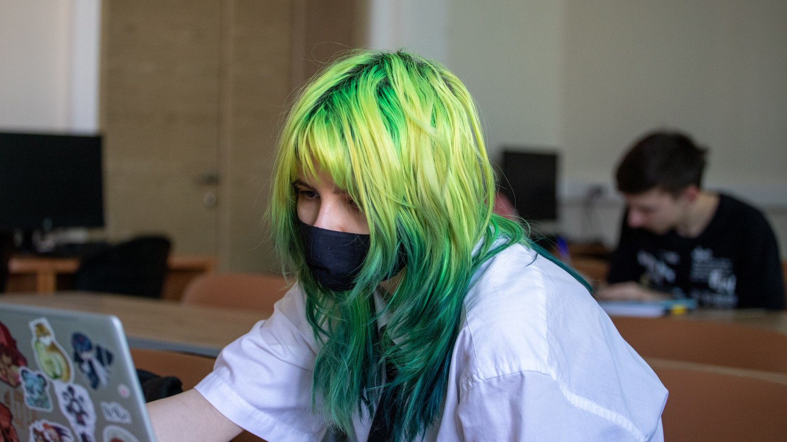 High school student with dyed green hair uses a laptop wearing a mask in classroom setting.
