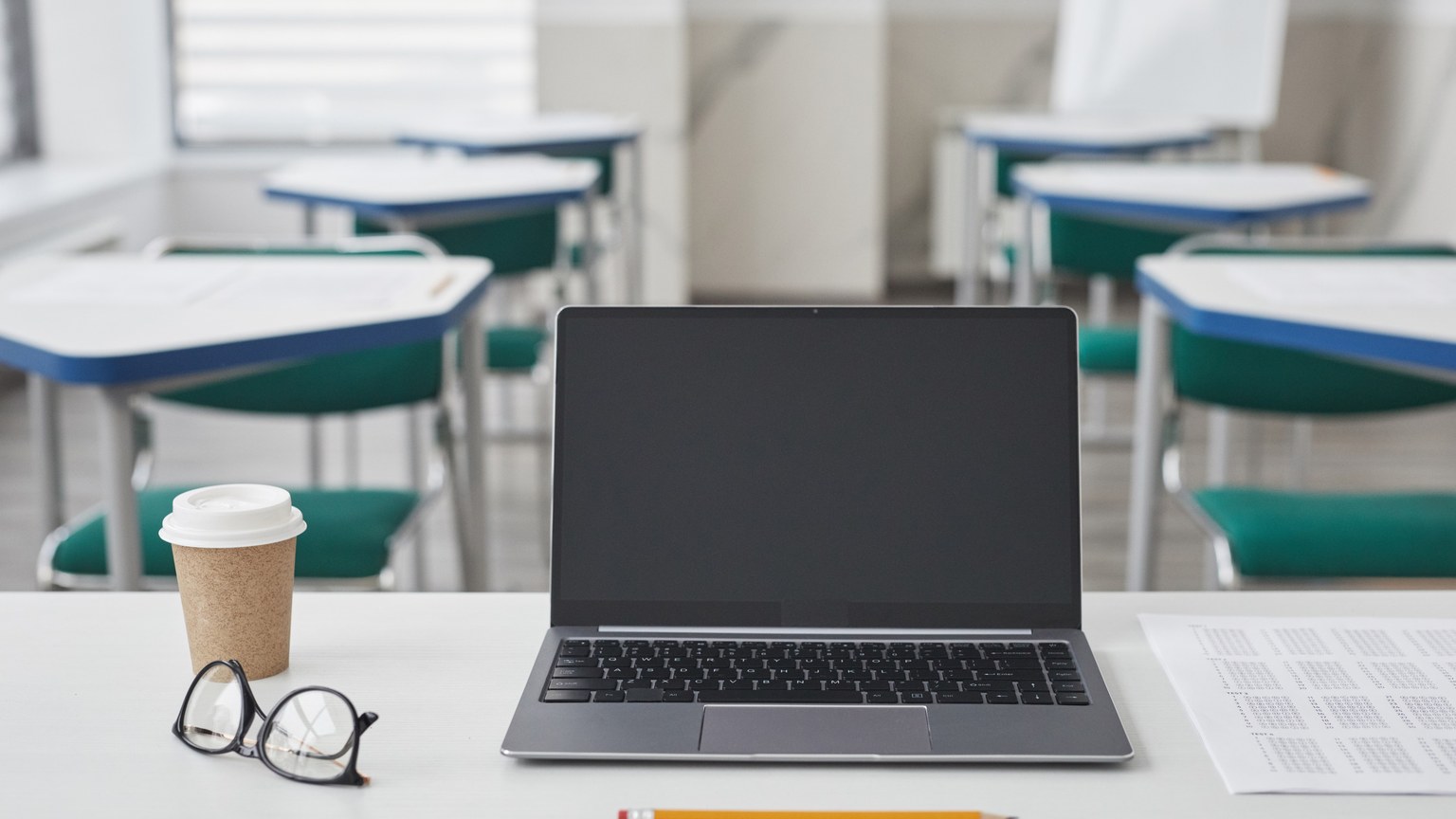 A neat classroom desk with a laptop, coffee cup, pencil, glasses, and paper.