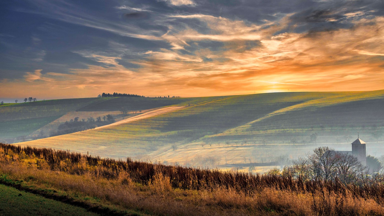 Breathtaking sunrise over rolling hills with fields and rural landscape, featuring mist and vibrant colors.