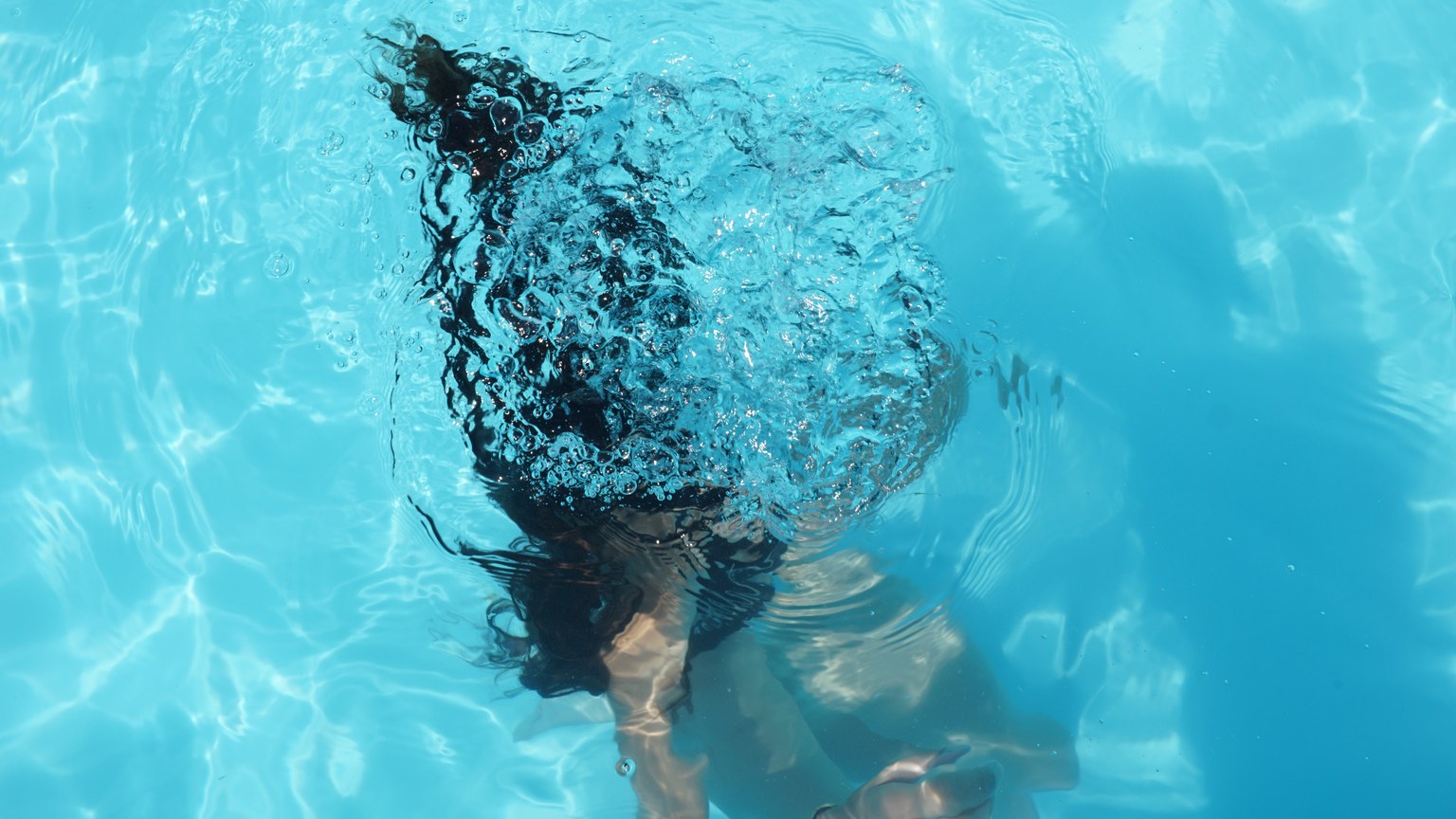 A woman diving underwater in a clear blue swimming pool, captured from above.