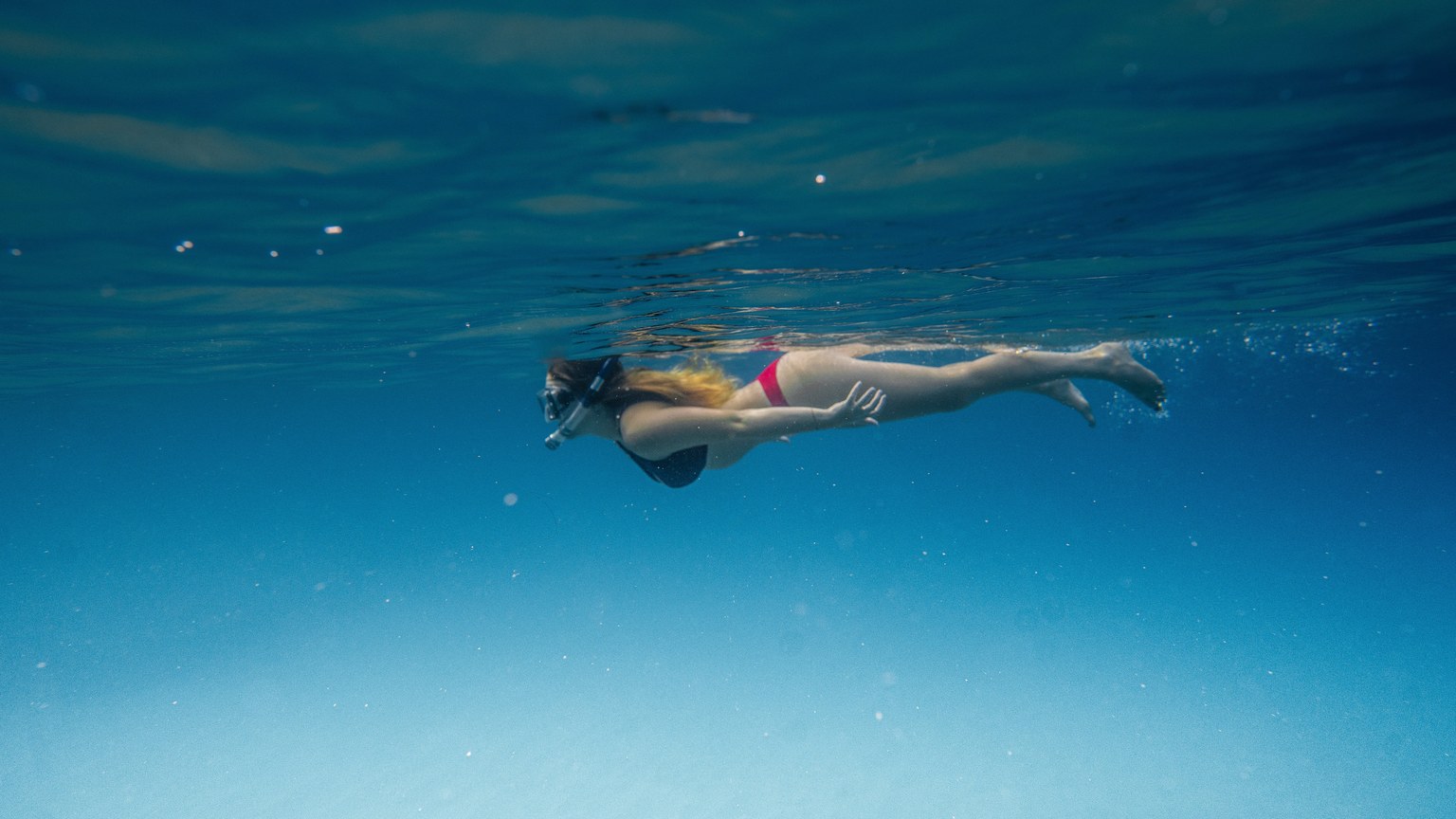 Side view of anonymous female swimmer in bikini and snorkeling mask diving in blue sea on sunny day