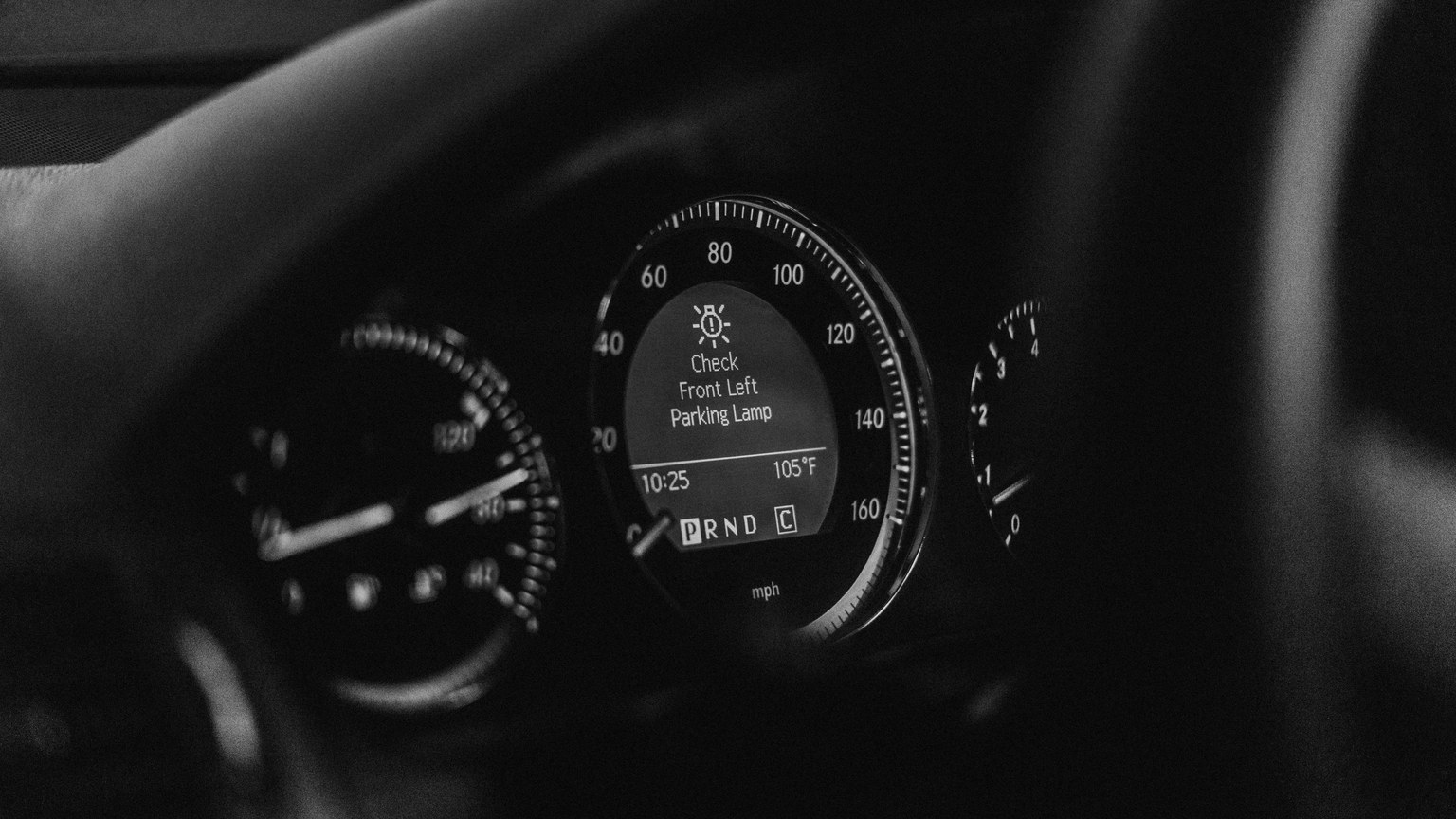 Close-up of a car dashboard in black and white with a parking lamp warning light.