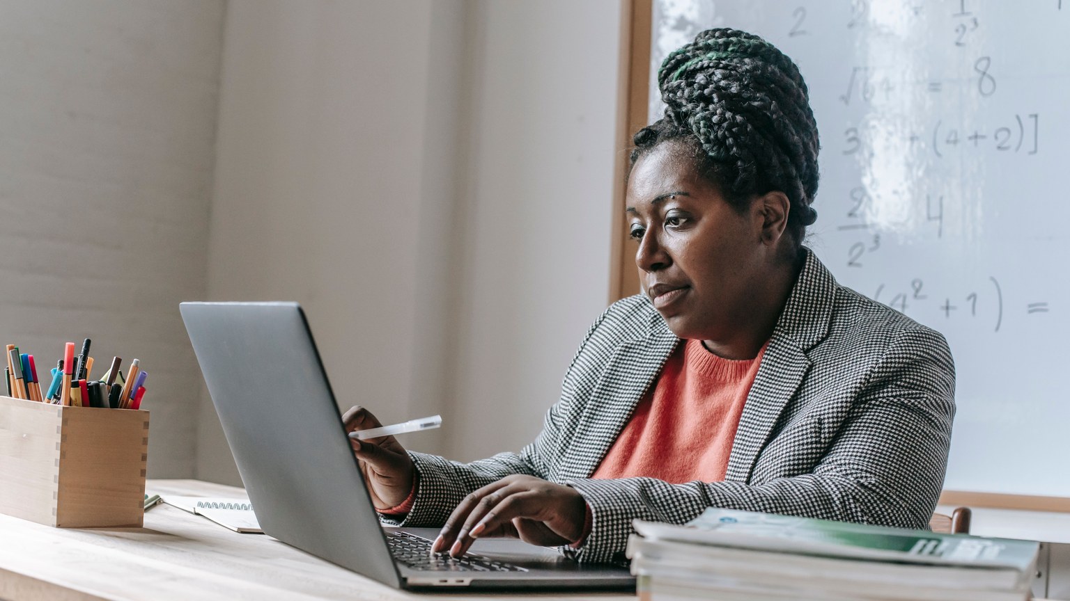 African American female teacher in formal outfit sitting at table in classroom and typing on laptop and taking notes on notepad