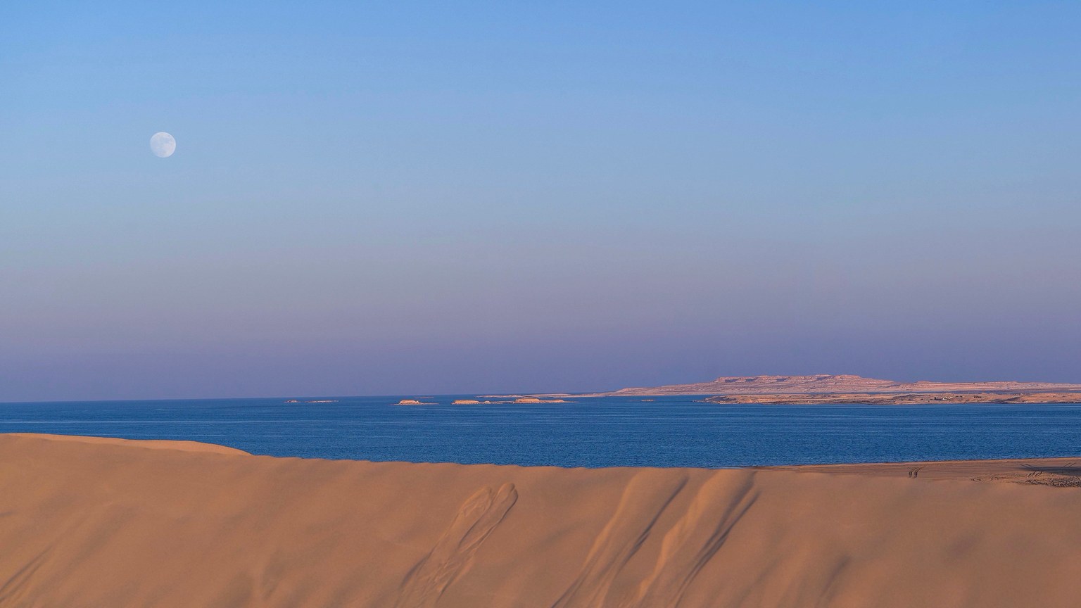 Amazing scenery of blue waving sea surrounded by sands of desert against cloudless sunset sky