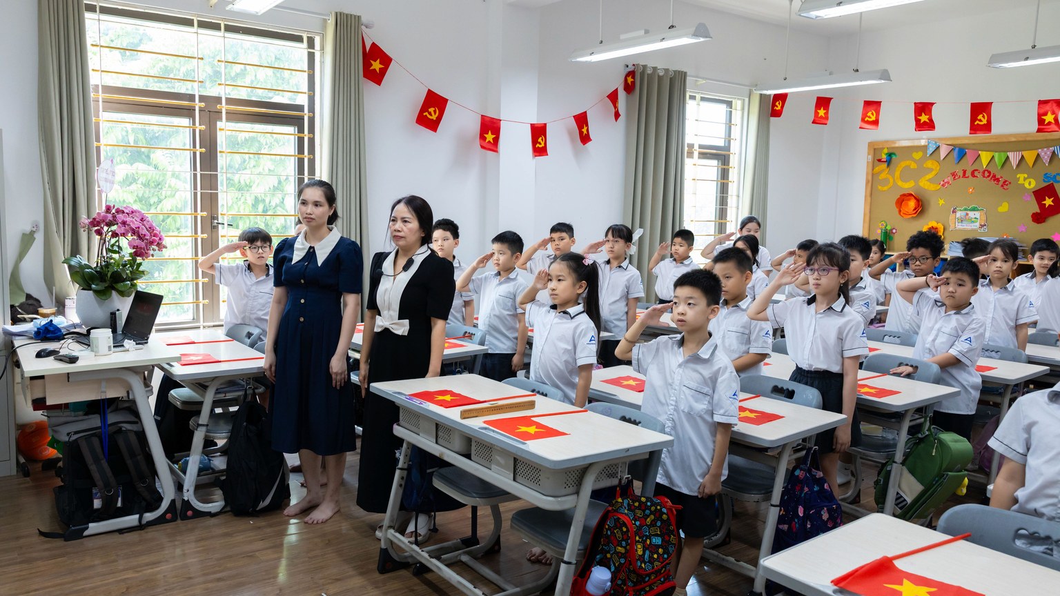 Students and teachers in a classroom standing and saluting with flags displayed.