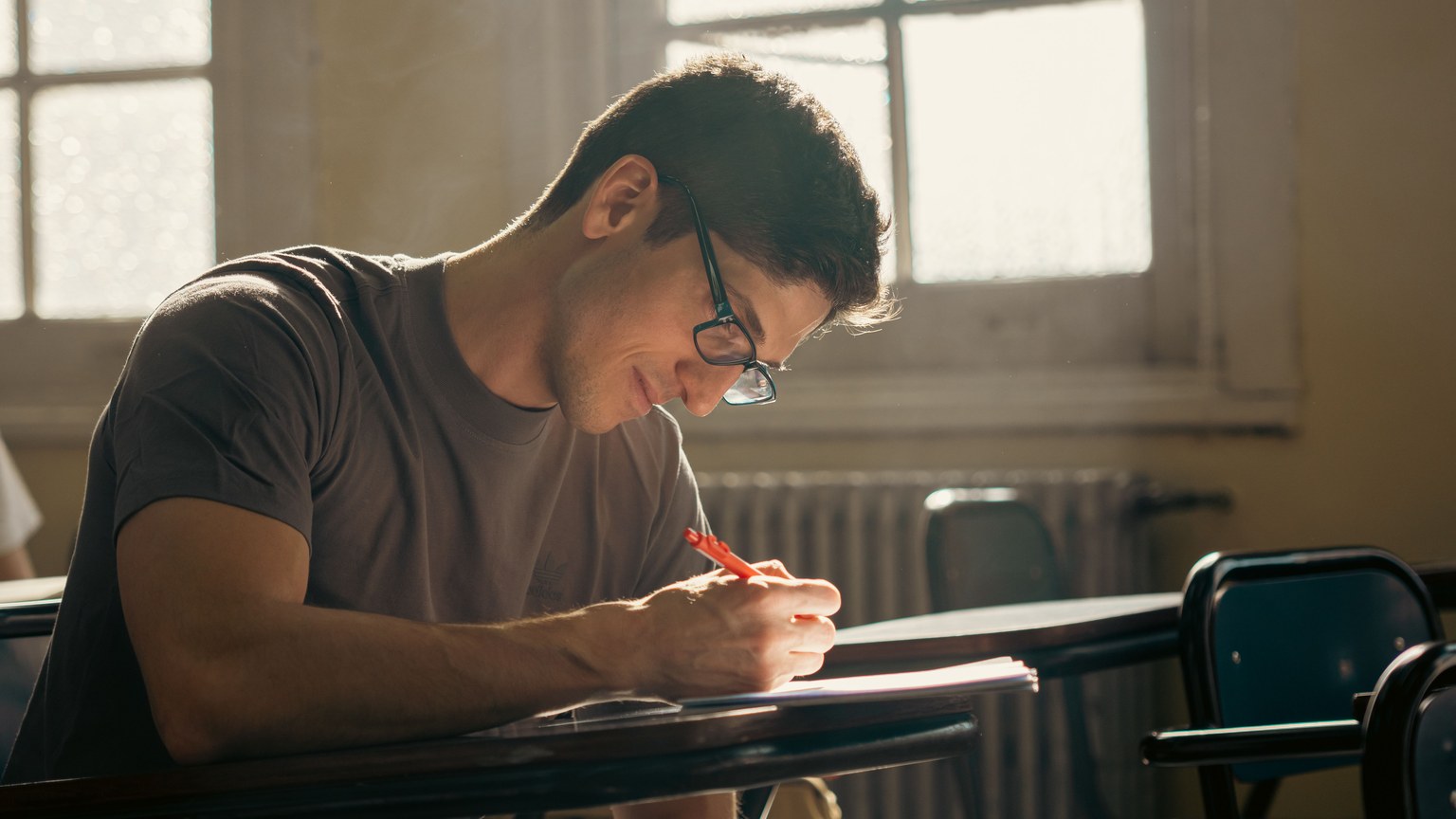 A student concentrating on writing during a class session in a warmly lit university classroom.
