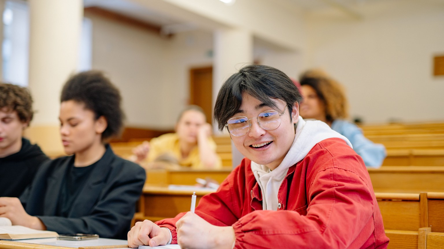Cheerful student wearing glasses seated in a university lecture hall takes notes during a class.