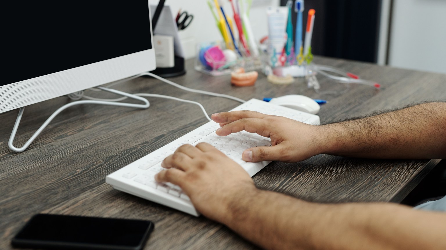 Close-up of hands typing on a white keyboard at a wooden desk in an office setting with accessories.
