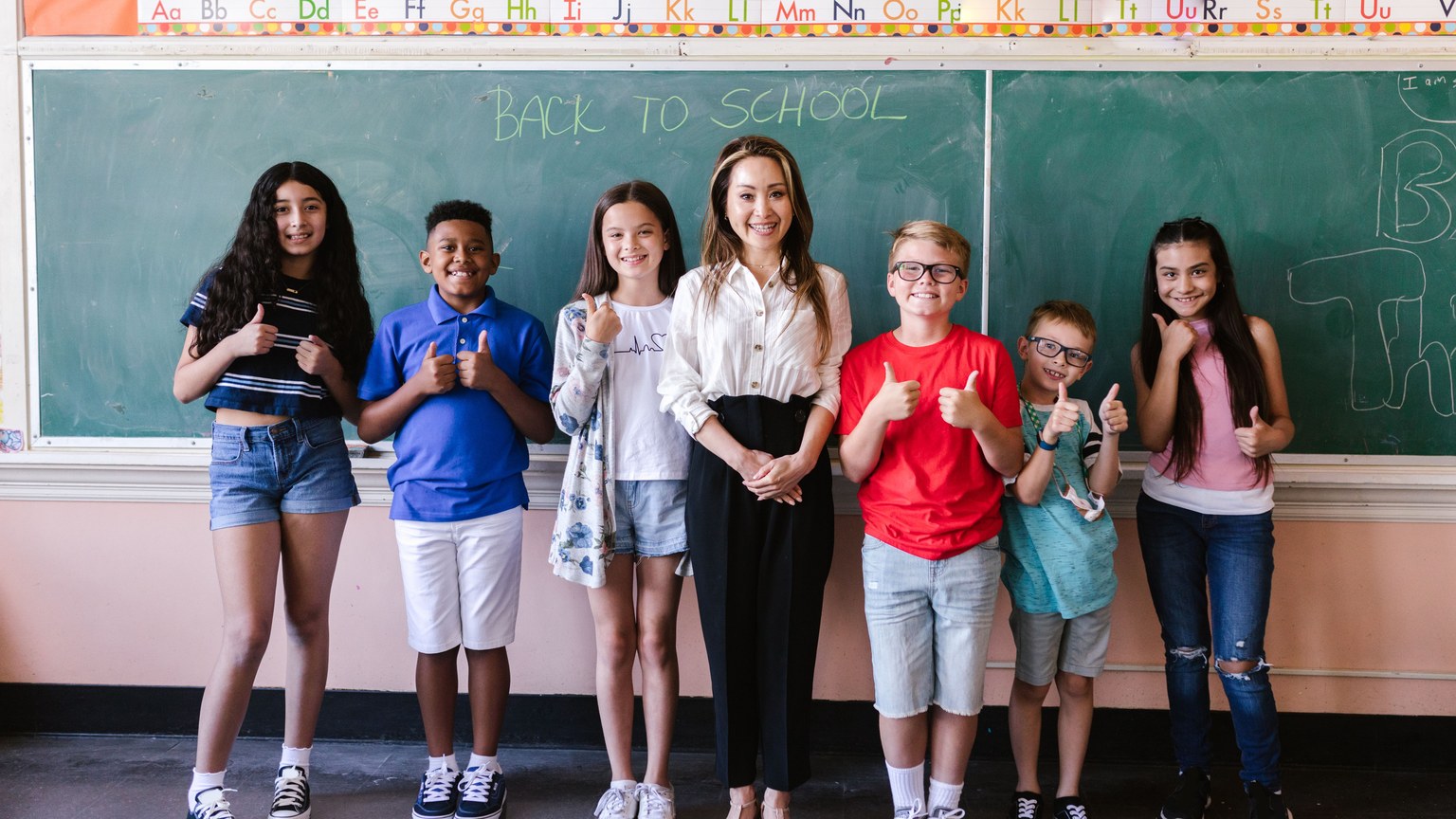 A diverse group of students and a teacher stand smiling in front of a back-to-school chalkboard.