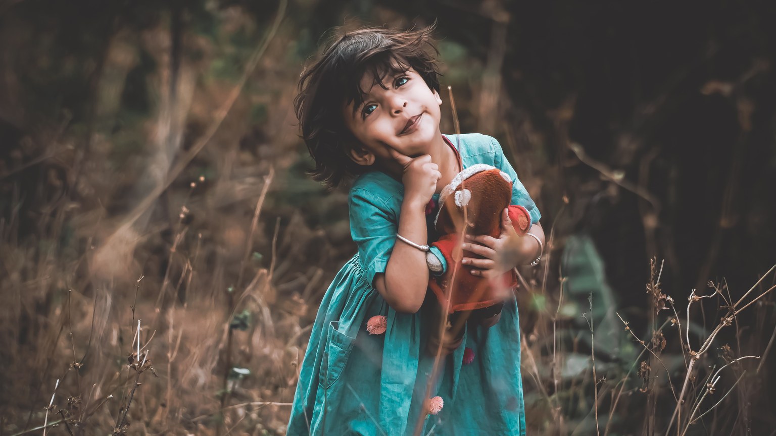 Charming young girl in a teal dress cuddling a toy, enjoying nature outdoors.
