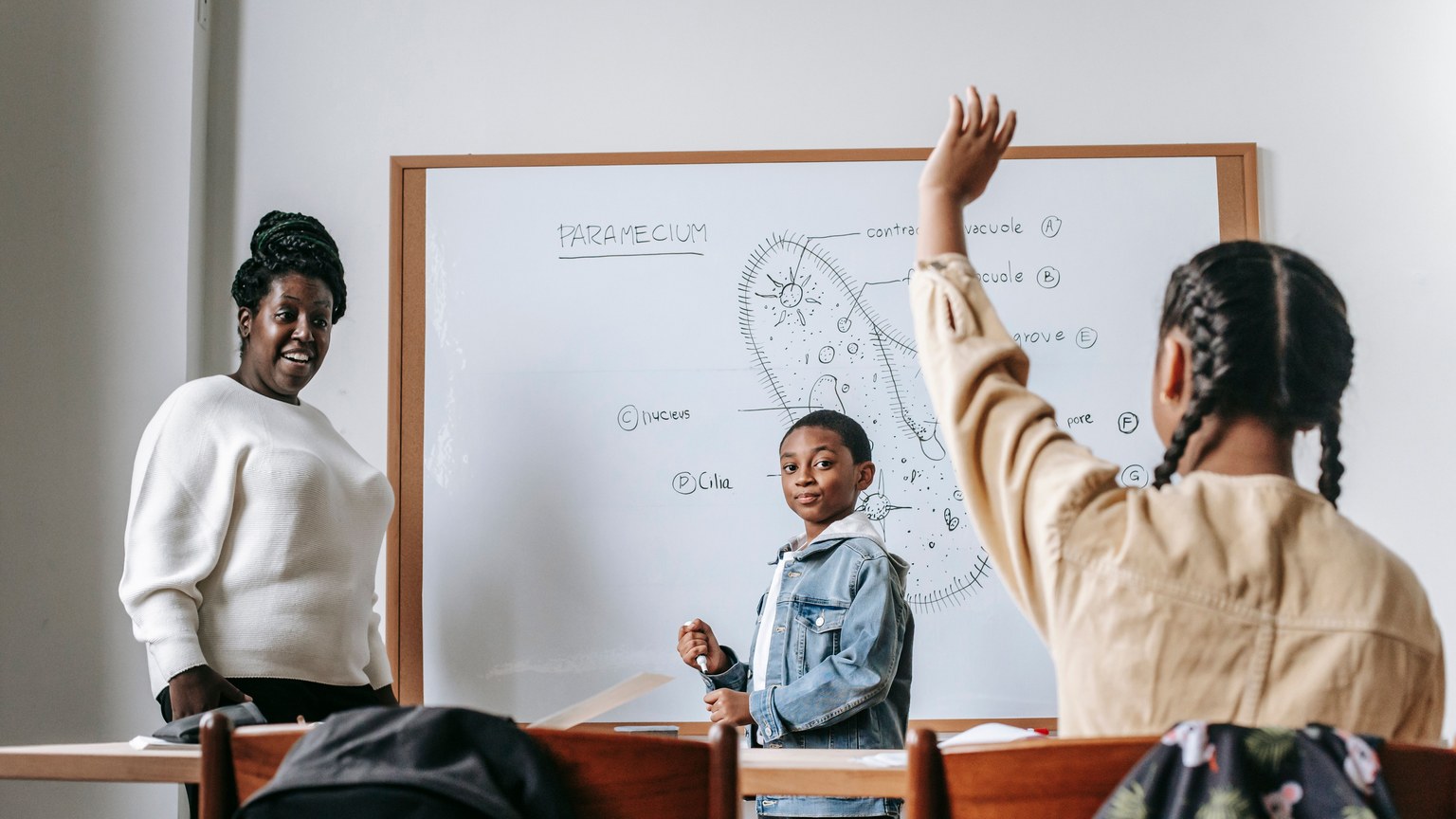 Ethnic girl raising hand while African American female teacher standing near whiteboard with teen boy and explaining task