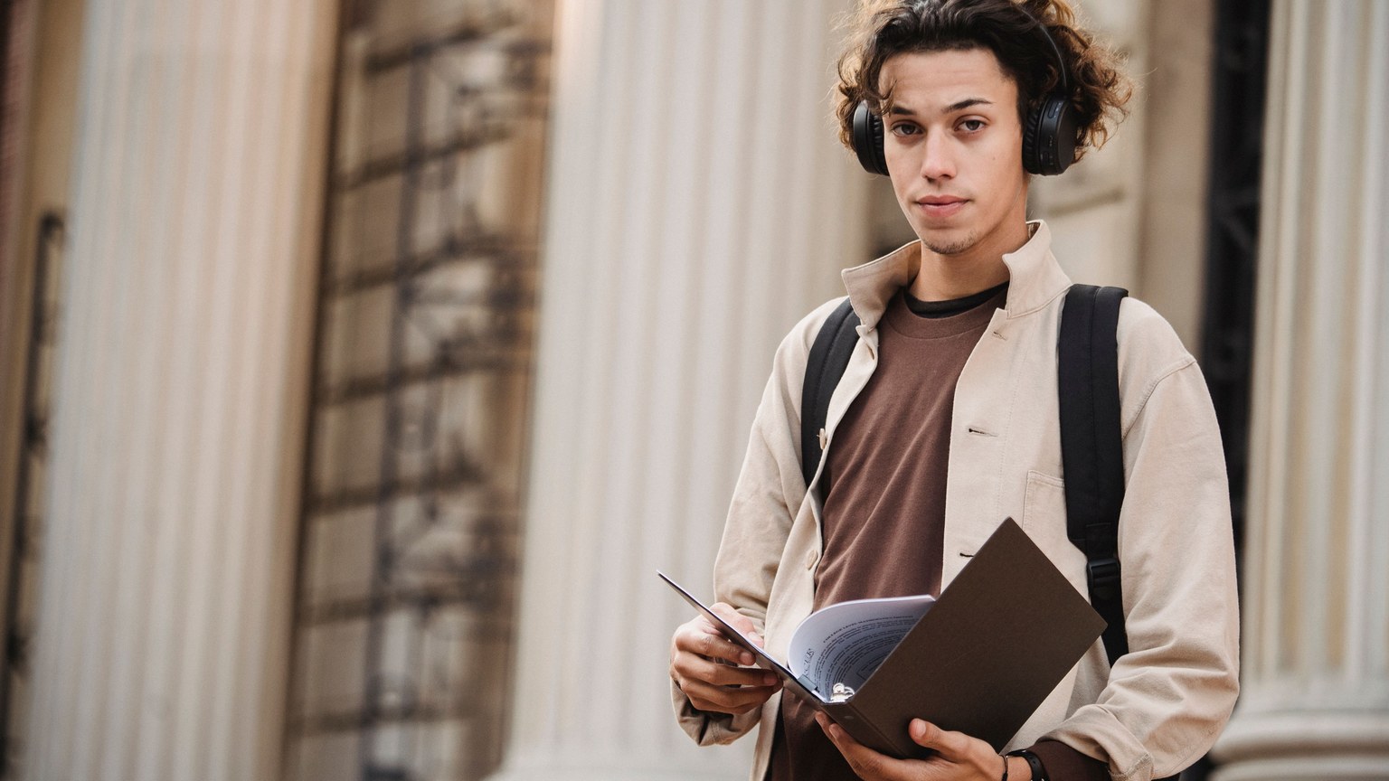 Young learner in wireless headphones in backpack standing with documents near entrance of university building