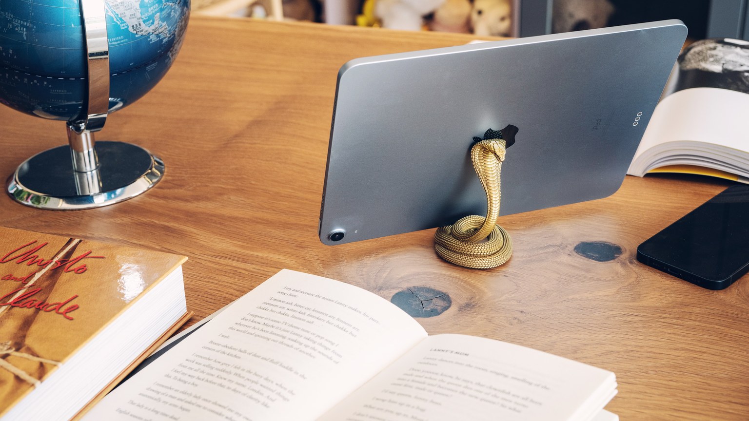 A snake-shaped brass stand holding a tablet on a wooden desk with books and a globe.