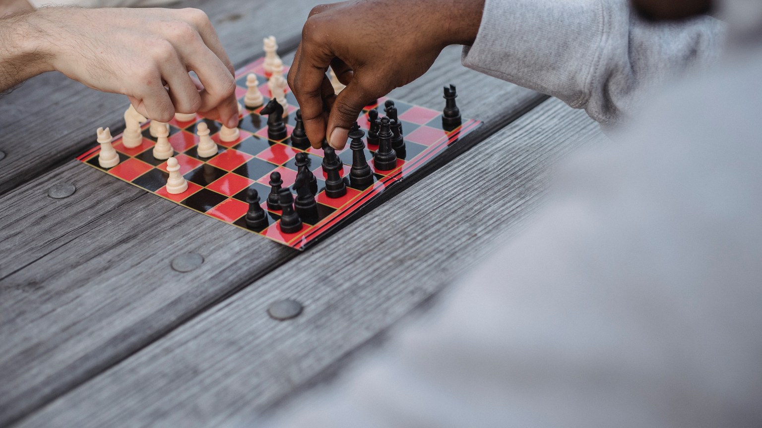High angle of crop faceless young diverse male friends playing chess sitting at wooden table in daylight