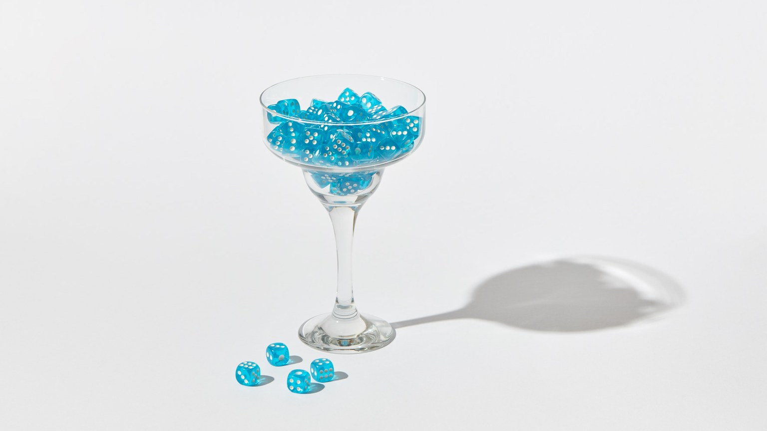 A cocktail glass filled with blue dice casting a shadow on a white background.