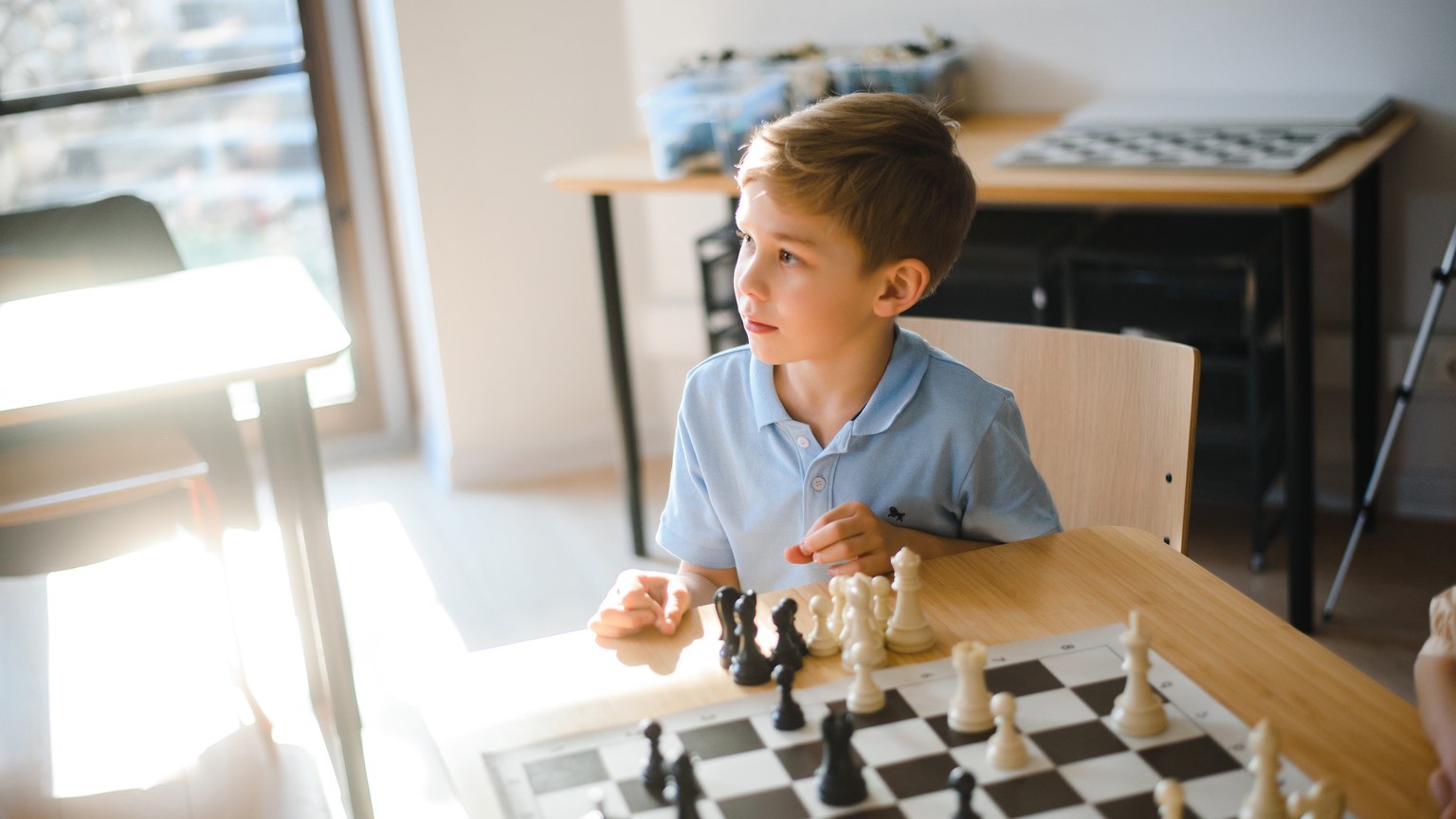 Boy in a blue shirt intently playing chess at a wooden table in a bright room.