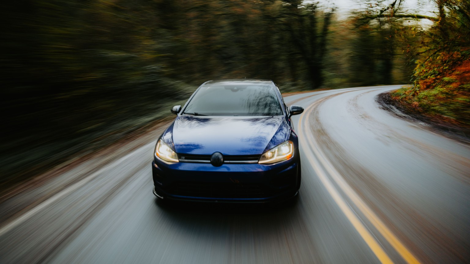 Dynamic shot of a blue car driving on a winding road through a scenic forest in autumn.