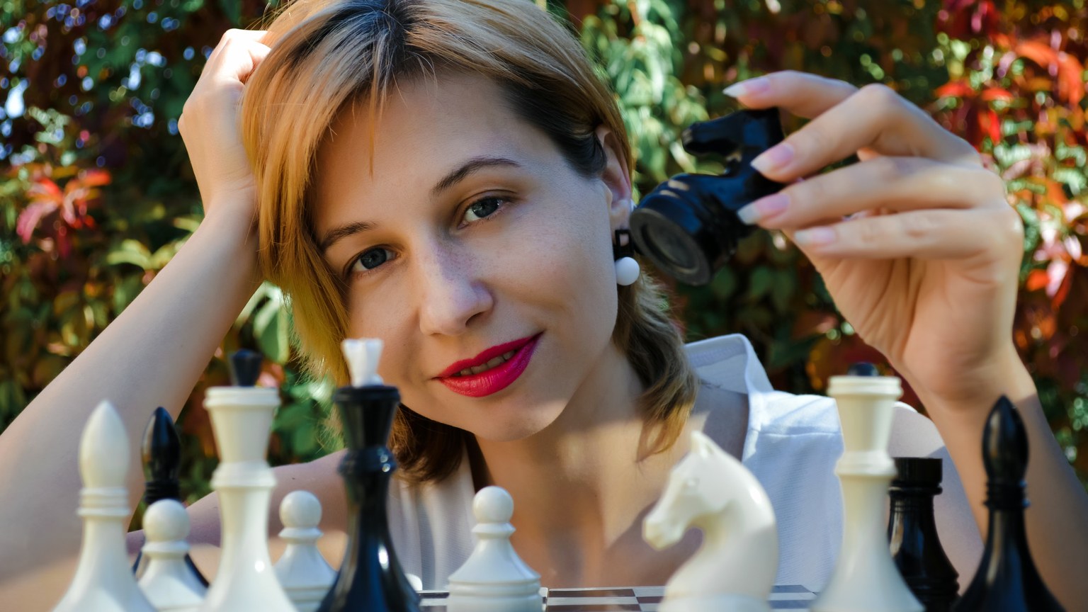 Glad young female chess player with small piece leaning on hand while sitting near board in park and looking at camera
