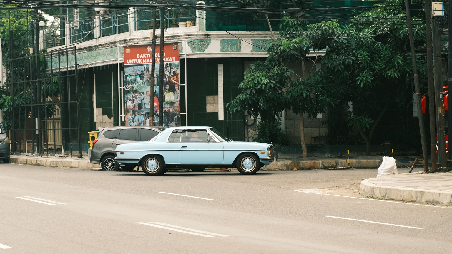 A classic light blue car parked on a tranquil street, backed by lush trees.