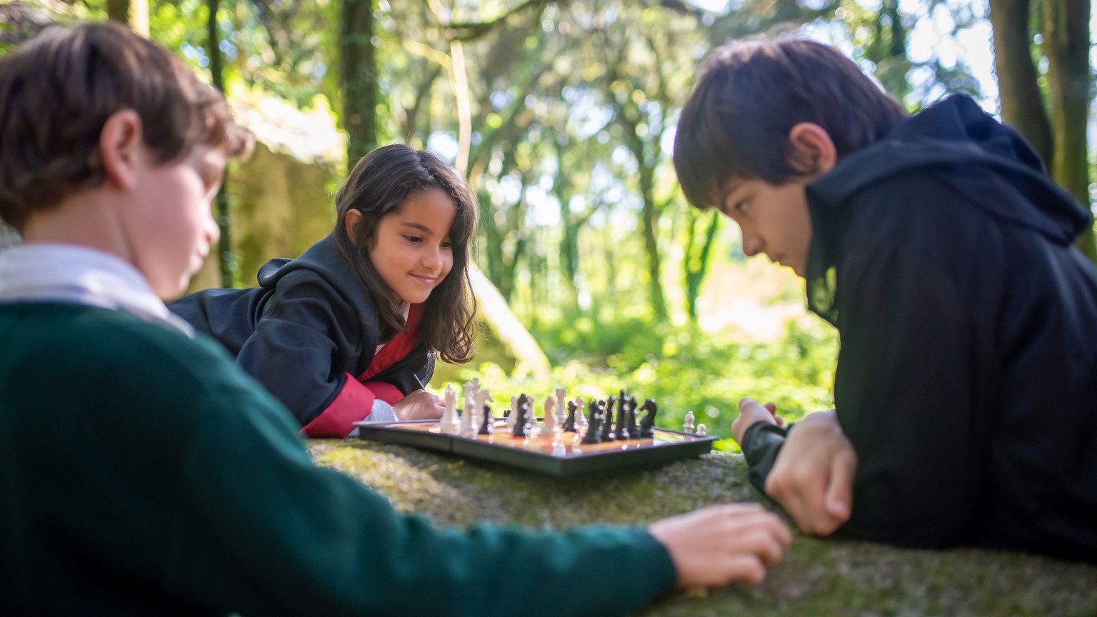 Three children in costumes playing chess on a stone outdoors in a forest.